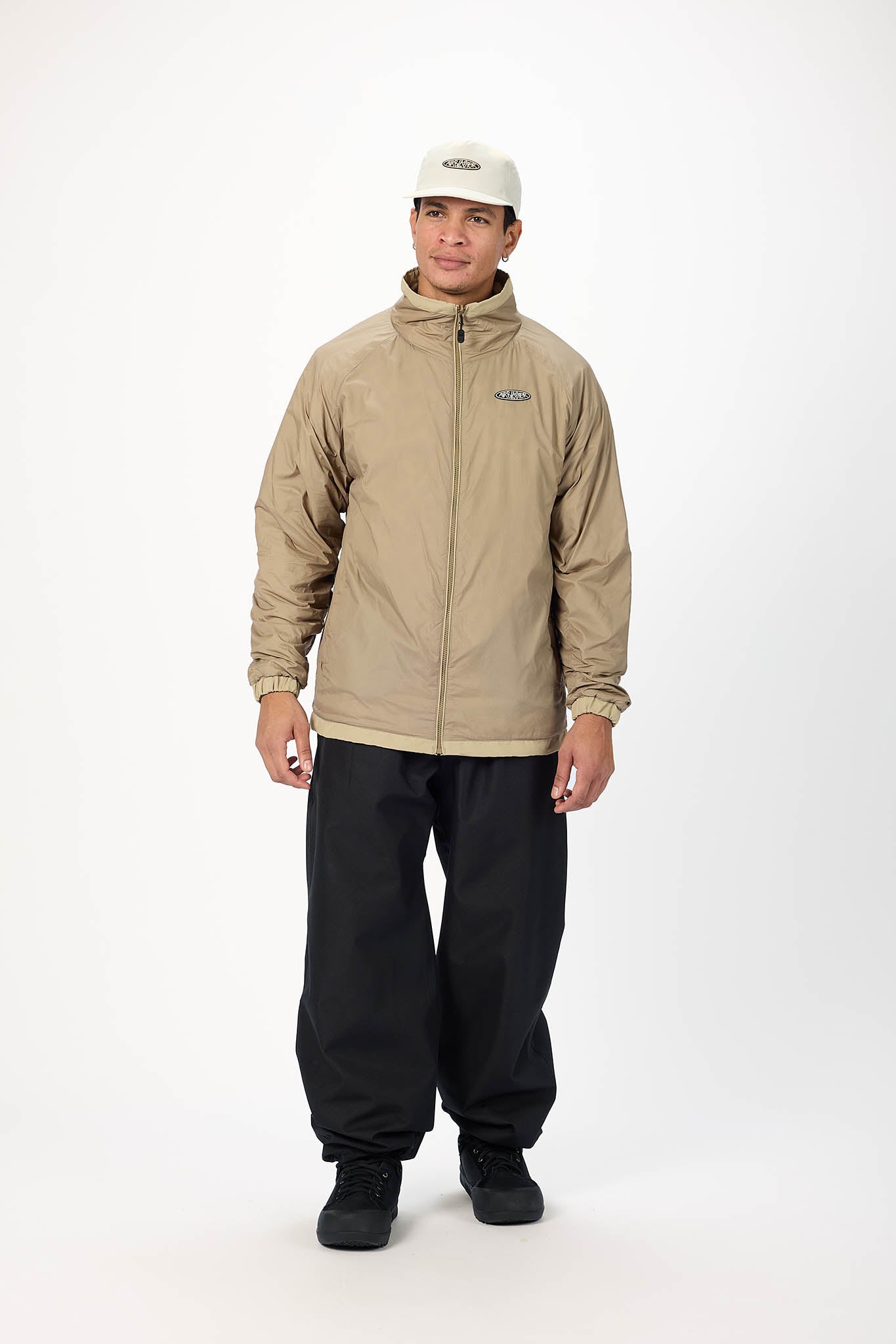 A man modeling a beige zip-up jacket with a high collar, black pants, and a white cap against a plain white studio background.
