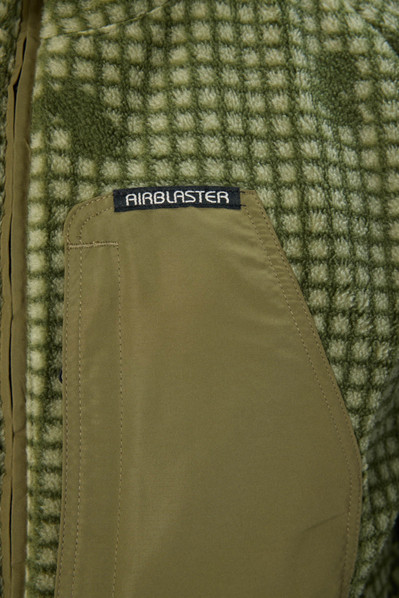 Close-up of a green and beige checkered fleece jacket with a tan zippered pocket and a black AIRBLASTER brand label.
