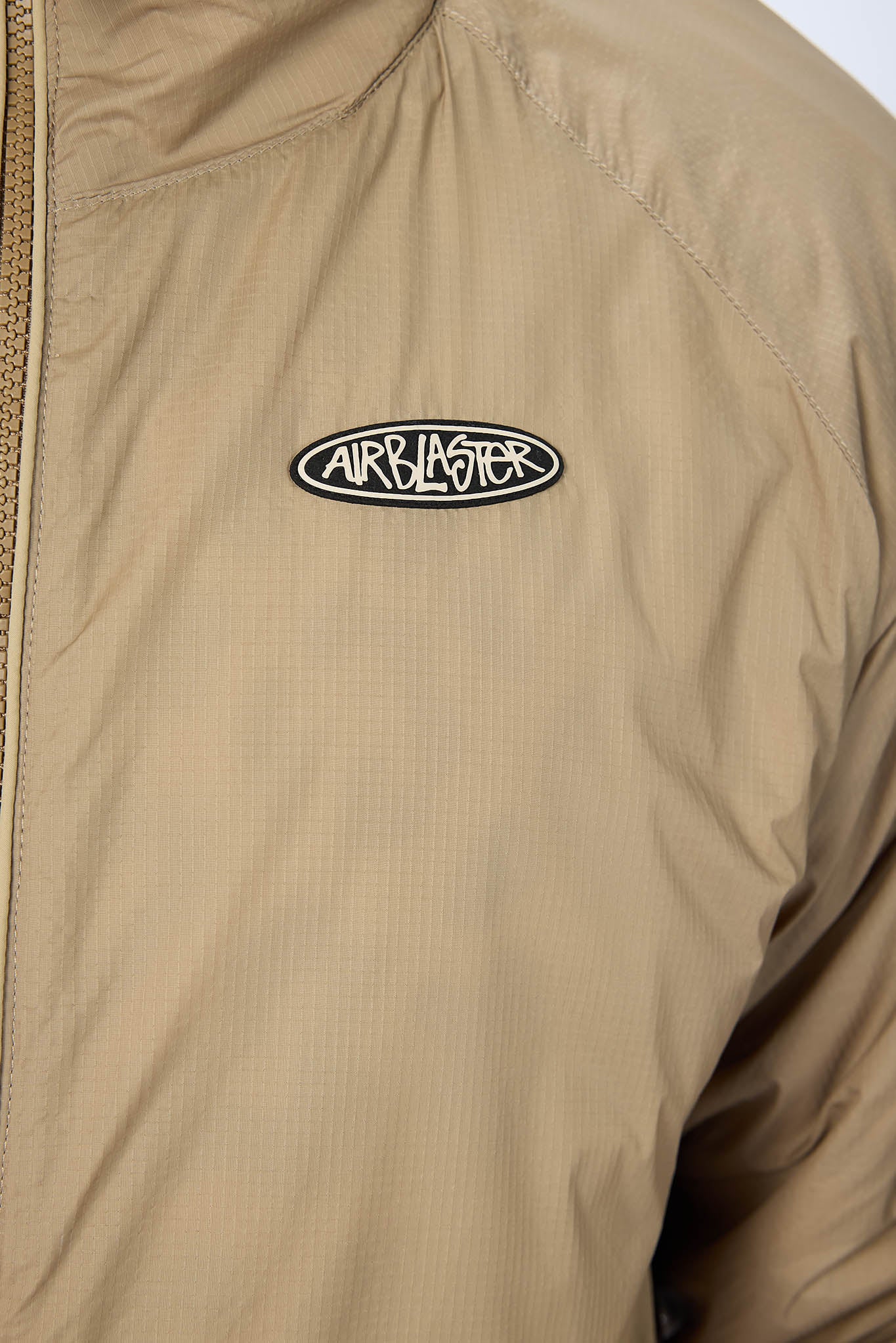 Close-up of a beige zip-front jacket with a black and white Airblaster logo patch on the chest.