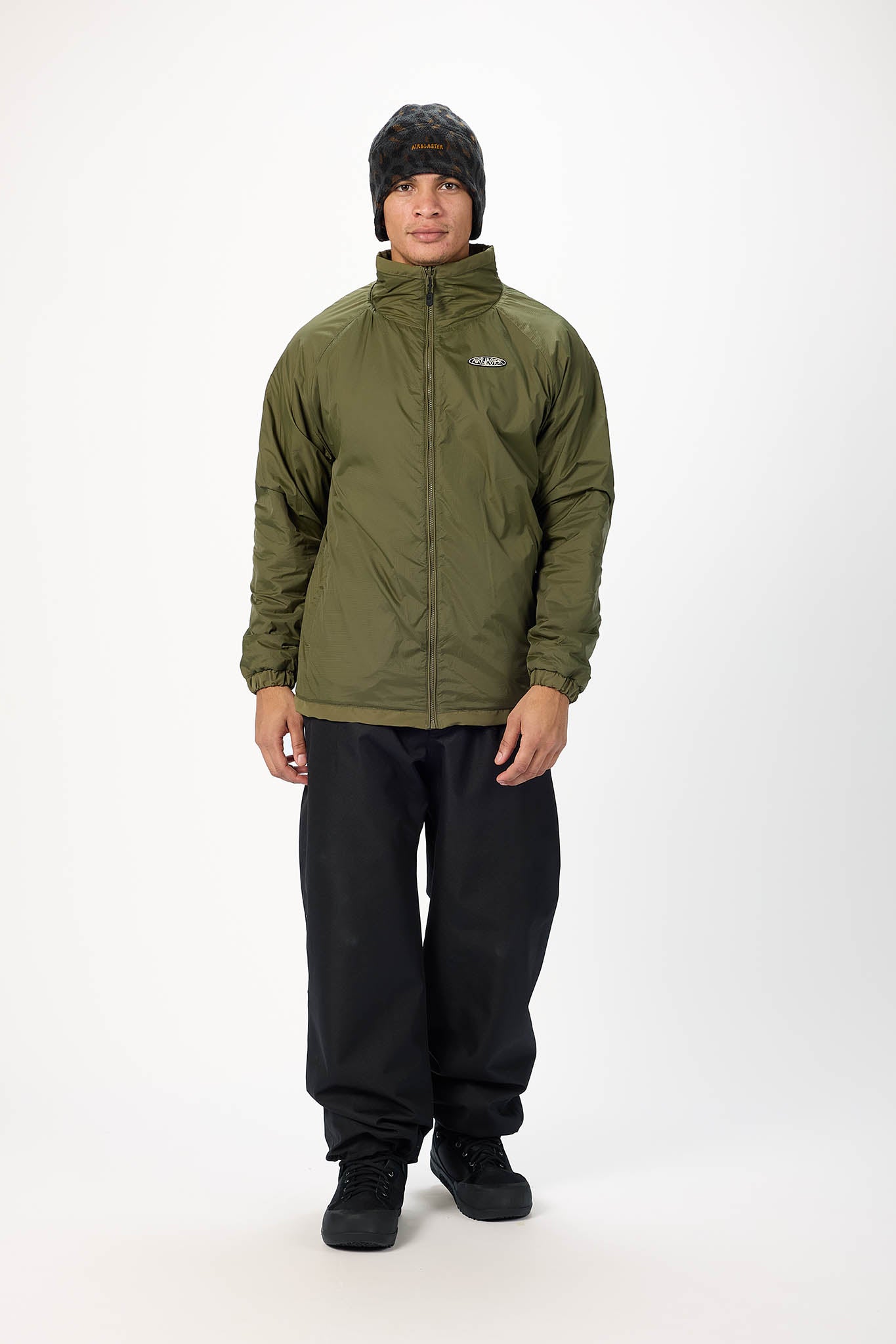 A man wearing an olive green zip-up jacket, black pants, and a black knit beanie stands against a plain white studio background.