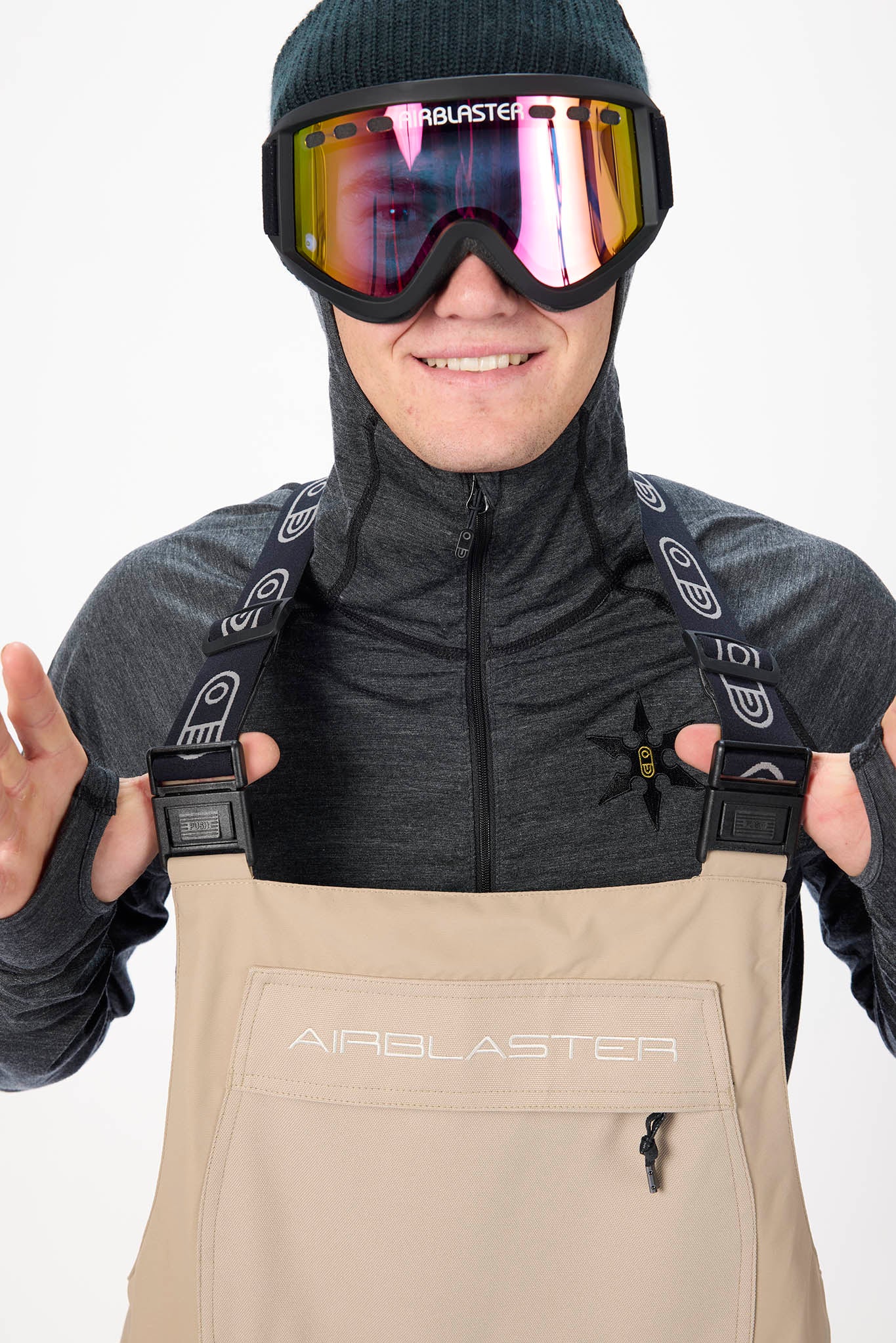 Person wearing a gray jacket, black helmet with goggles, and beige bib overalls with 'Airblaster' branding.