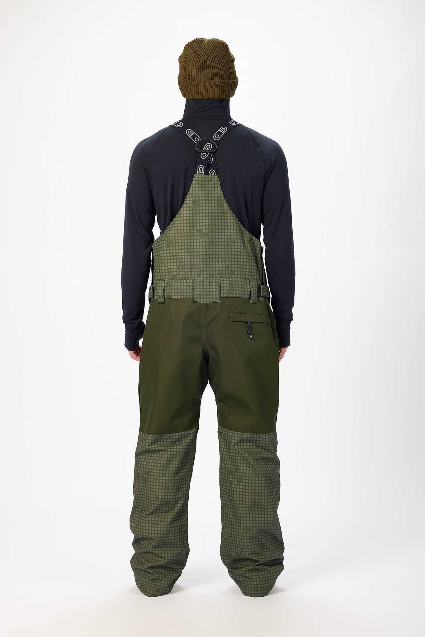 Person wearing a dark jacket, green bib overalls, and a matching beanie on a white background