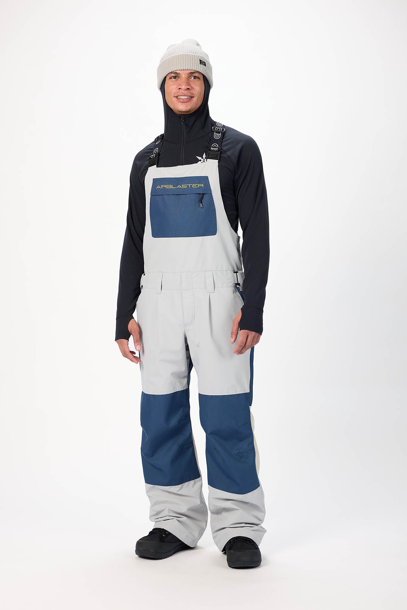 Person wearing a black jacket, gray bib overalls, and black boots on a white background