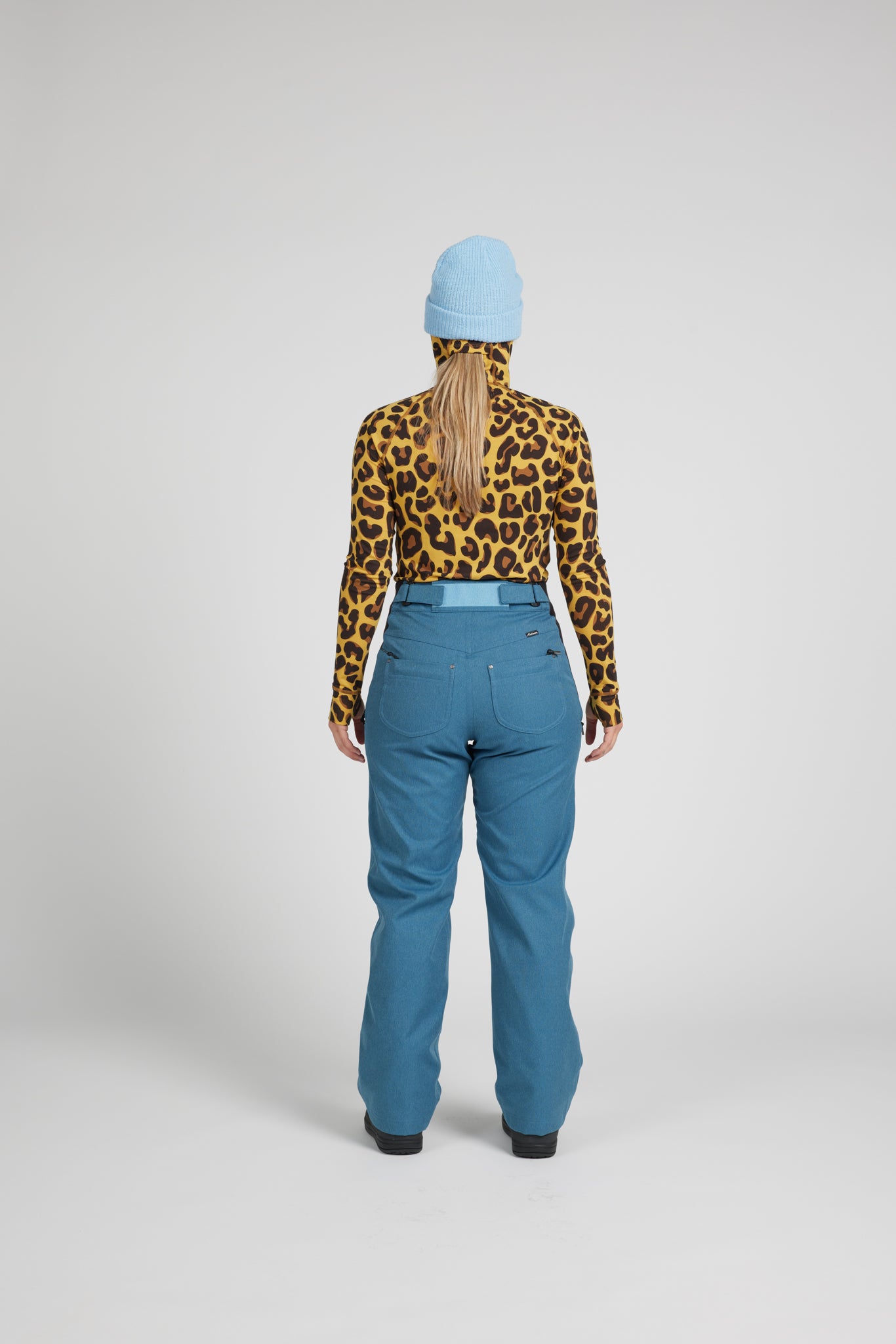 A model shown from the back wearing a yellow and black leopard print long-sleeve top, blue ski pants, a light blue beanie, and a light blue belt, against a light gray studio background.
