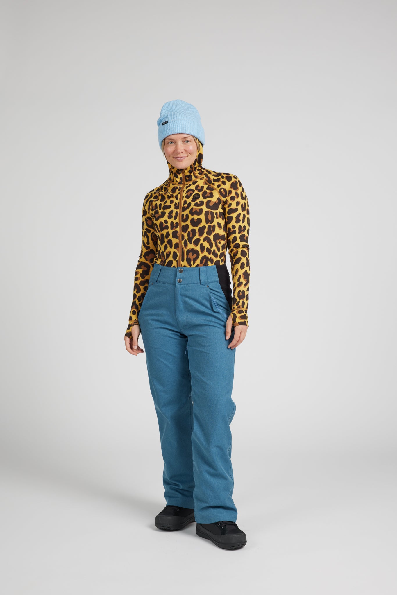 A model wearing a yellow and black leopard print mock neck top, teal blue ski pants, a light blue beanie, and black shoes, standing against a light gray studio background.