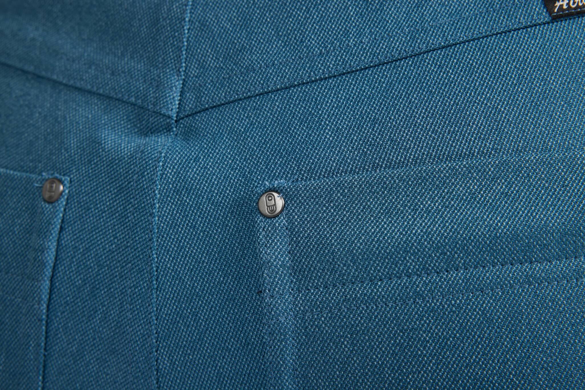 Close-up of a textured teal blue fabric garment, showing a pocket with a metal snap button and part of a dark brand label in the top right corner.