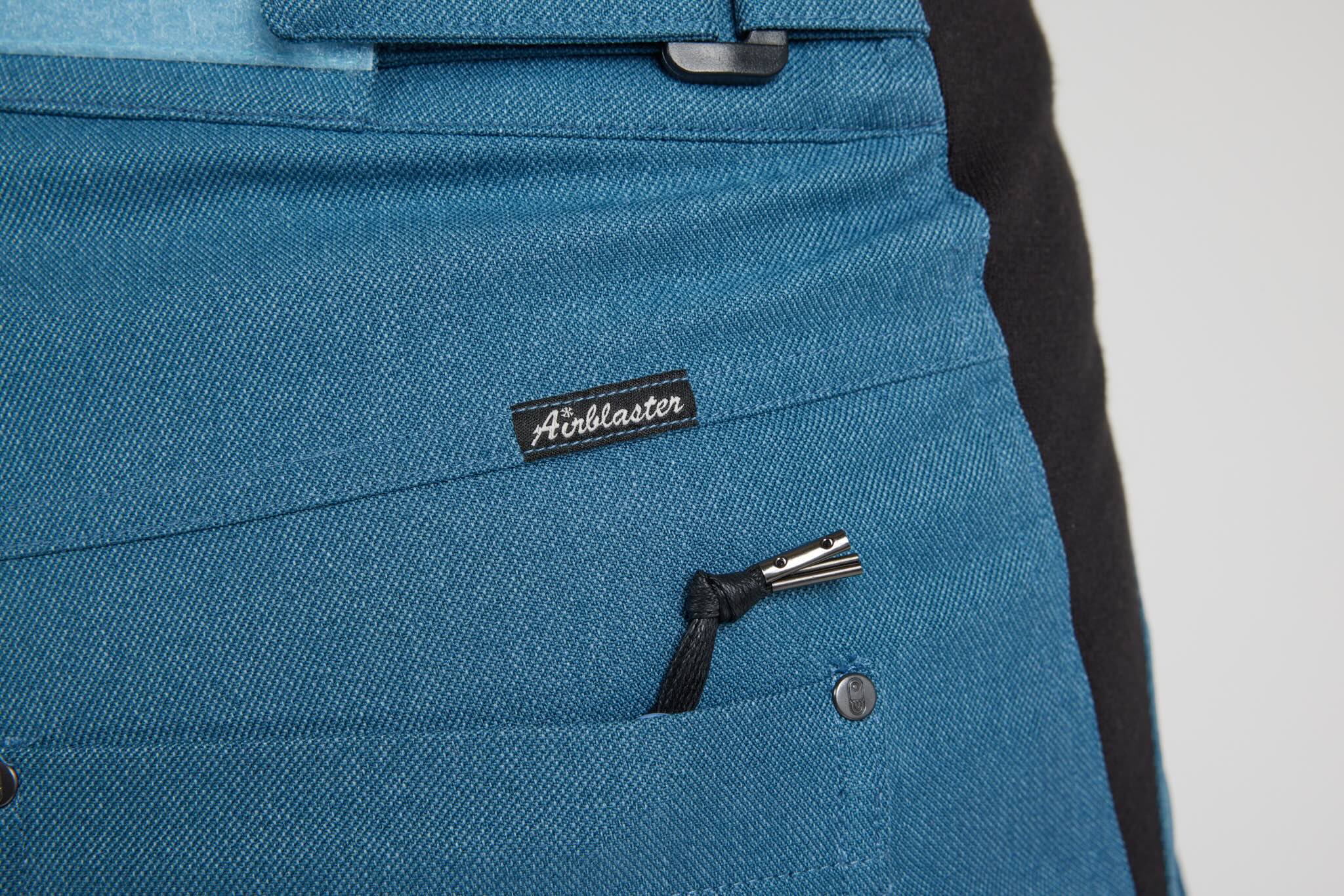 Close-up of a blue canvas work apron with a black brand label reading 'Airblaster' and a black drawstring zipper pocket.