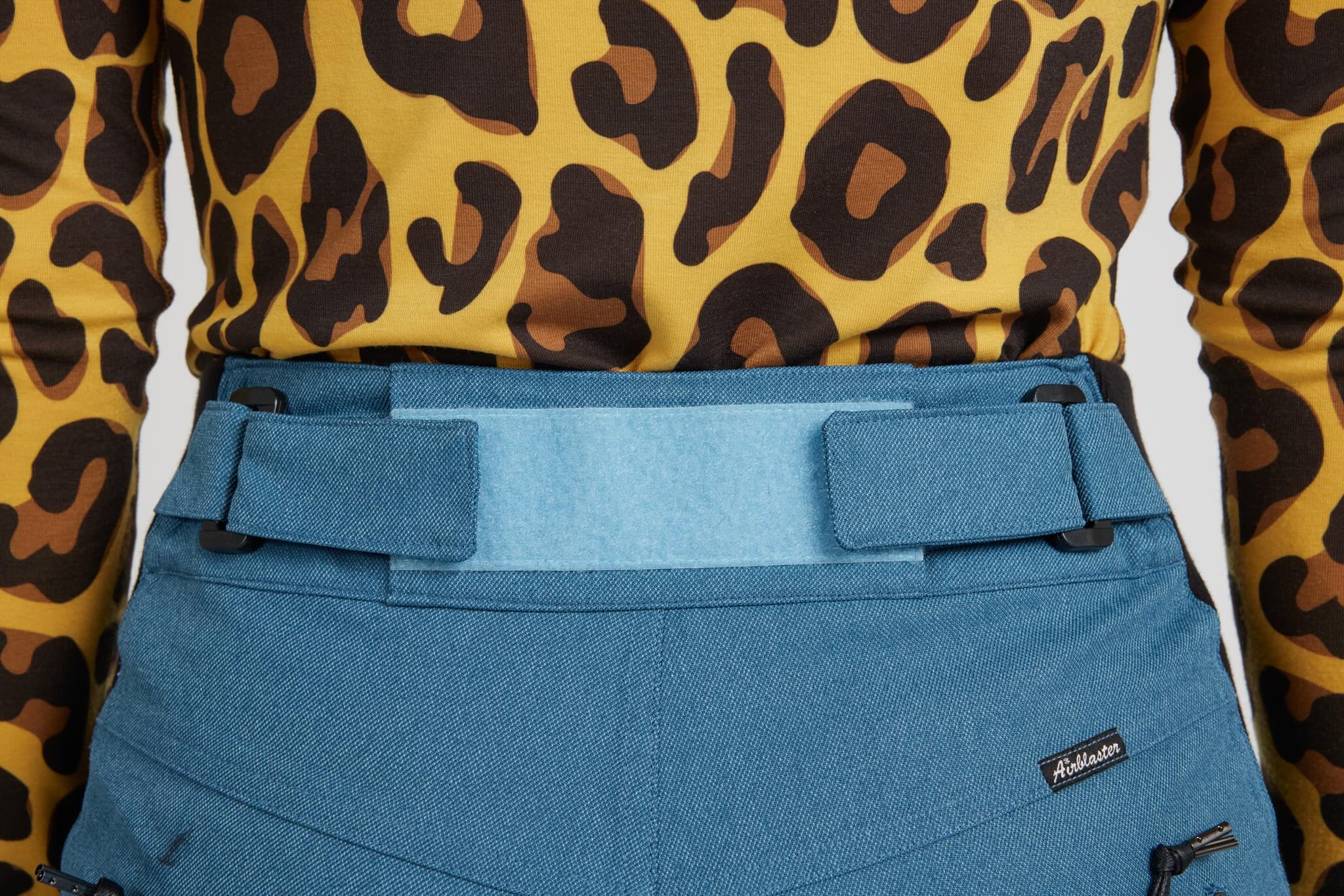 Close-up of a person wearing blue ski pants with a wide waist belt and a yellow leopard-print long-sleeve top. A small black brand tag reading 'Adidas' is visible on the pants.