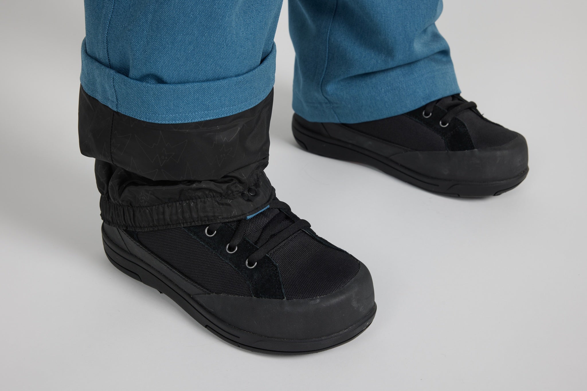 A close-up of a person's feet wearing black lace-up work boots with black ankle cuffs, paired with blue jeans, on a light gray studio background.