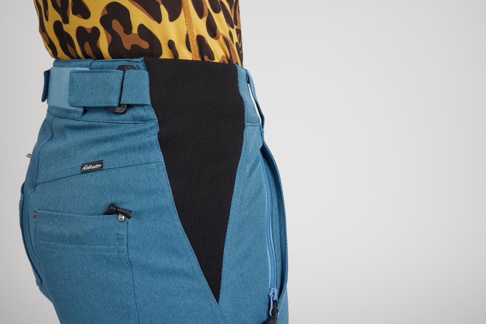 Close-up of a person wearing high-waisted blue trousers with a black center panel and a matching blue belt, paired with a leopard-print top against a light gray background.