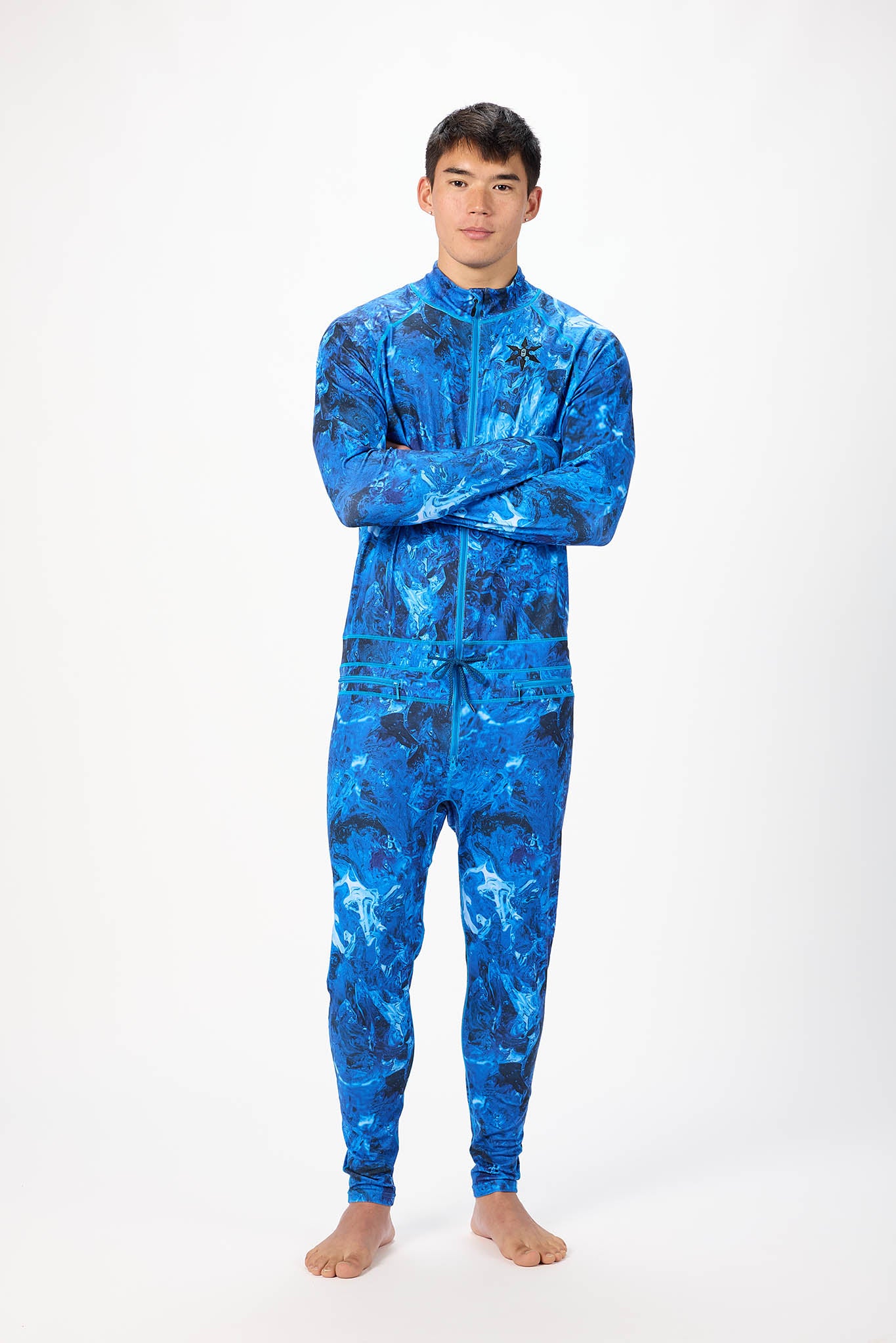 A man modeling a blue abstract pattern full-zip athletic onesie with a small white logo on the left chest, standing against a plain white studio background.