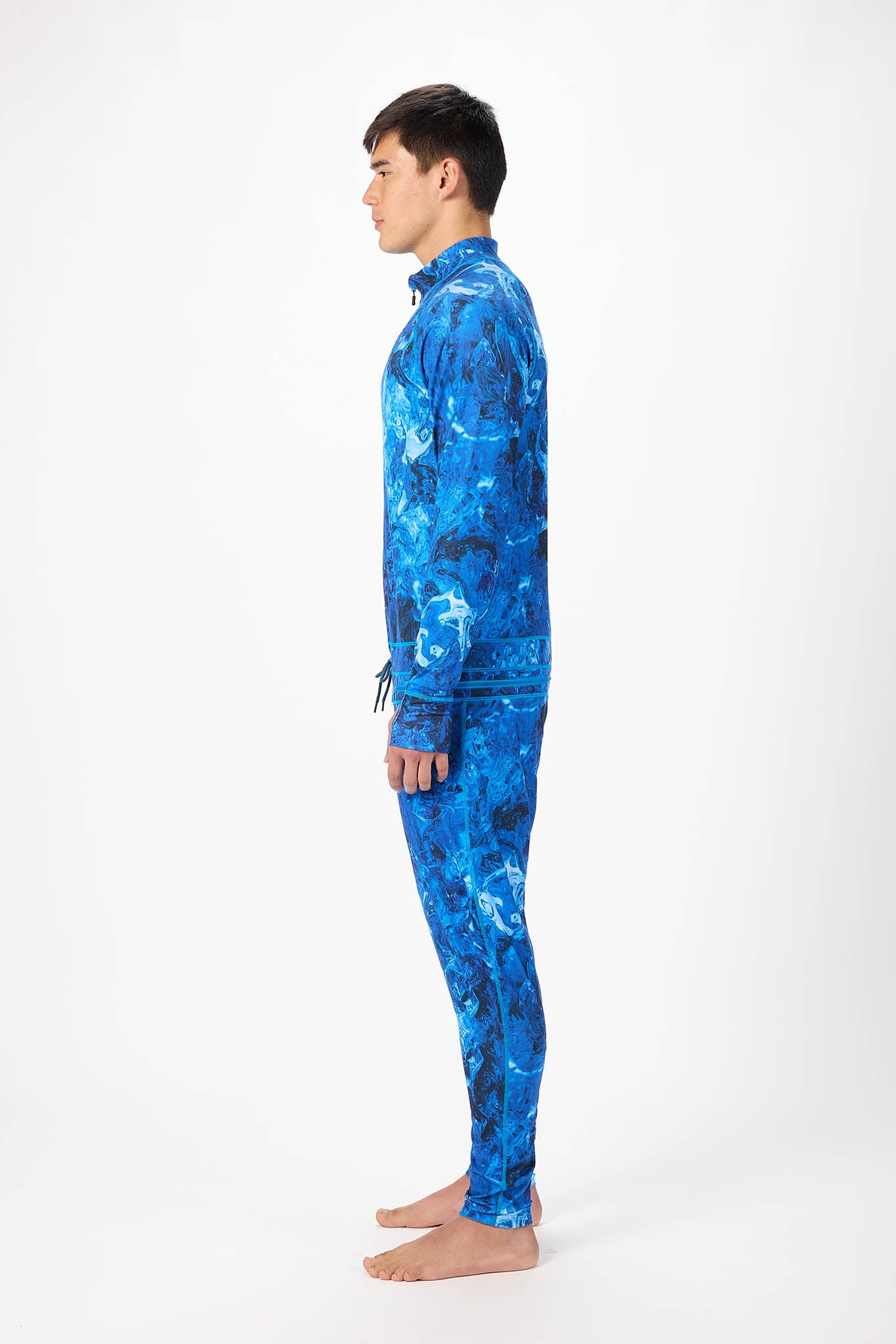 in side profile A man modeling a blue abstract pattern full-zip athletic onesie with a small white logo on the left chest, standing against a plain white studio background.