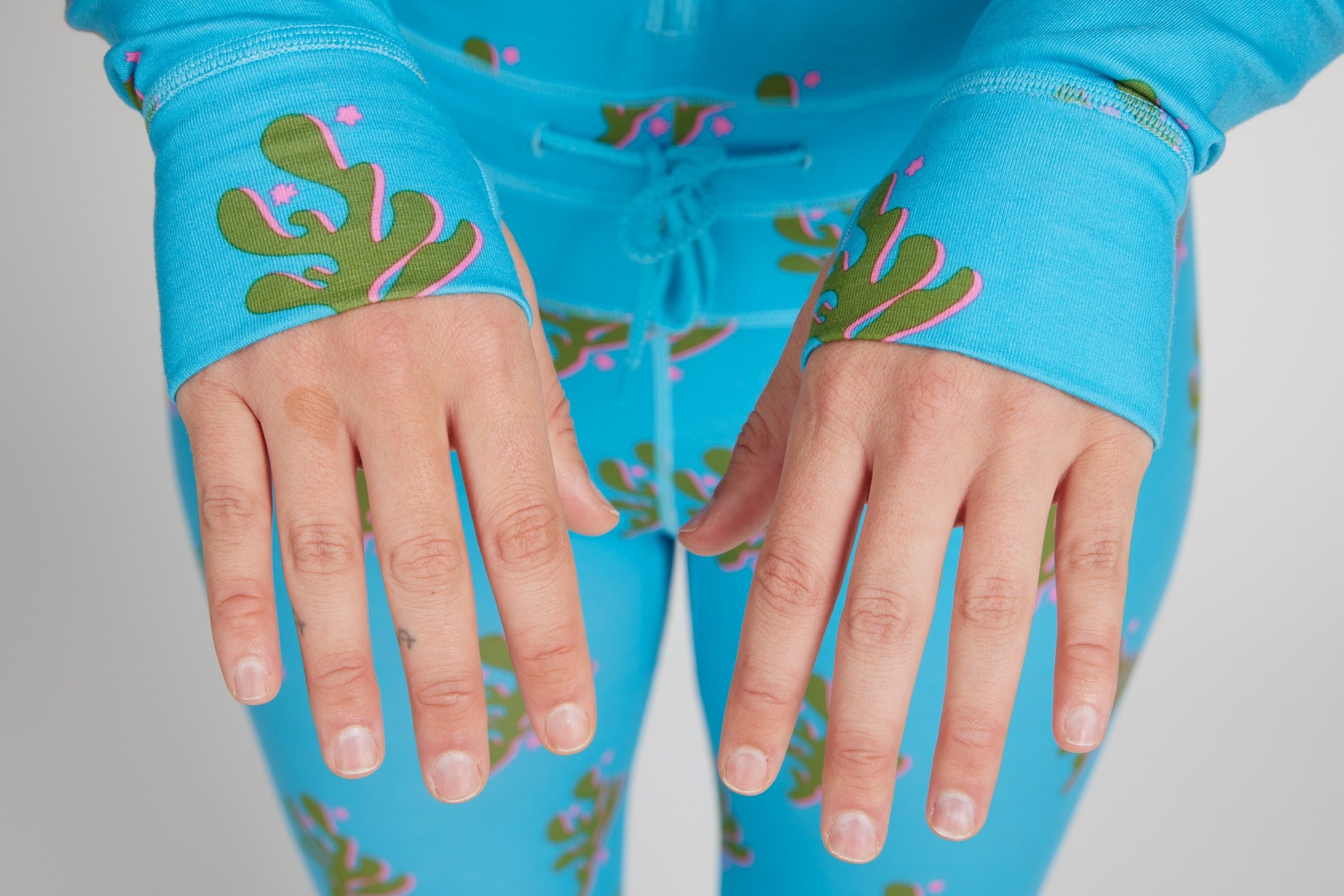 Close-up of a person wearing bright turquoise pajama pants and matching long-sleeve top with a green cactus print and pink accents, showing the cuffs and hands against a light gray background.