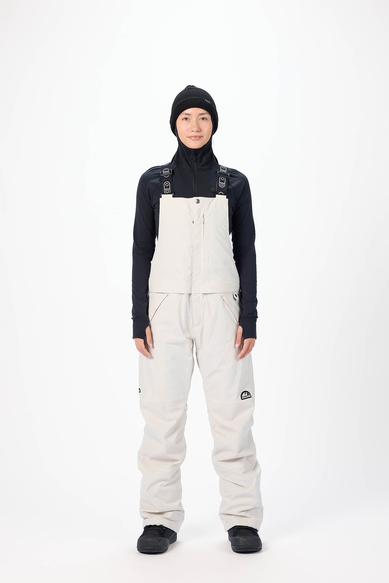 Person wearing a black jacket, white bib overalls, and black beanie on a white background