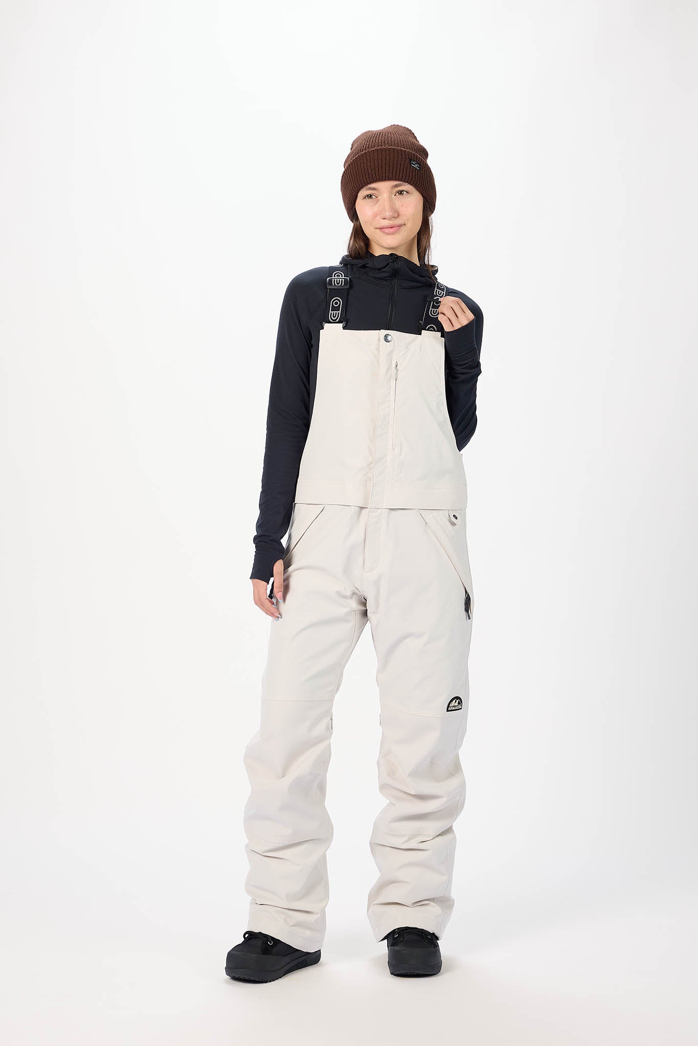 Person wearing a black jacket and beige bib overalls on a white background