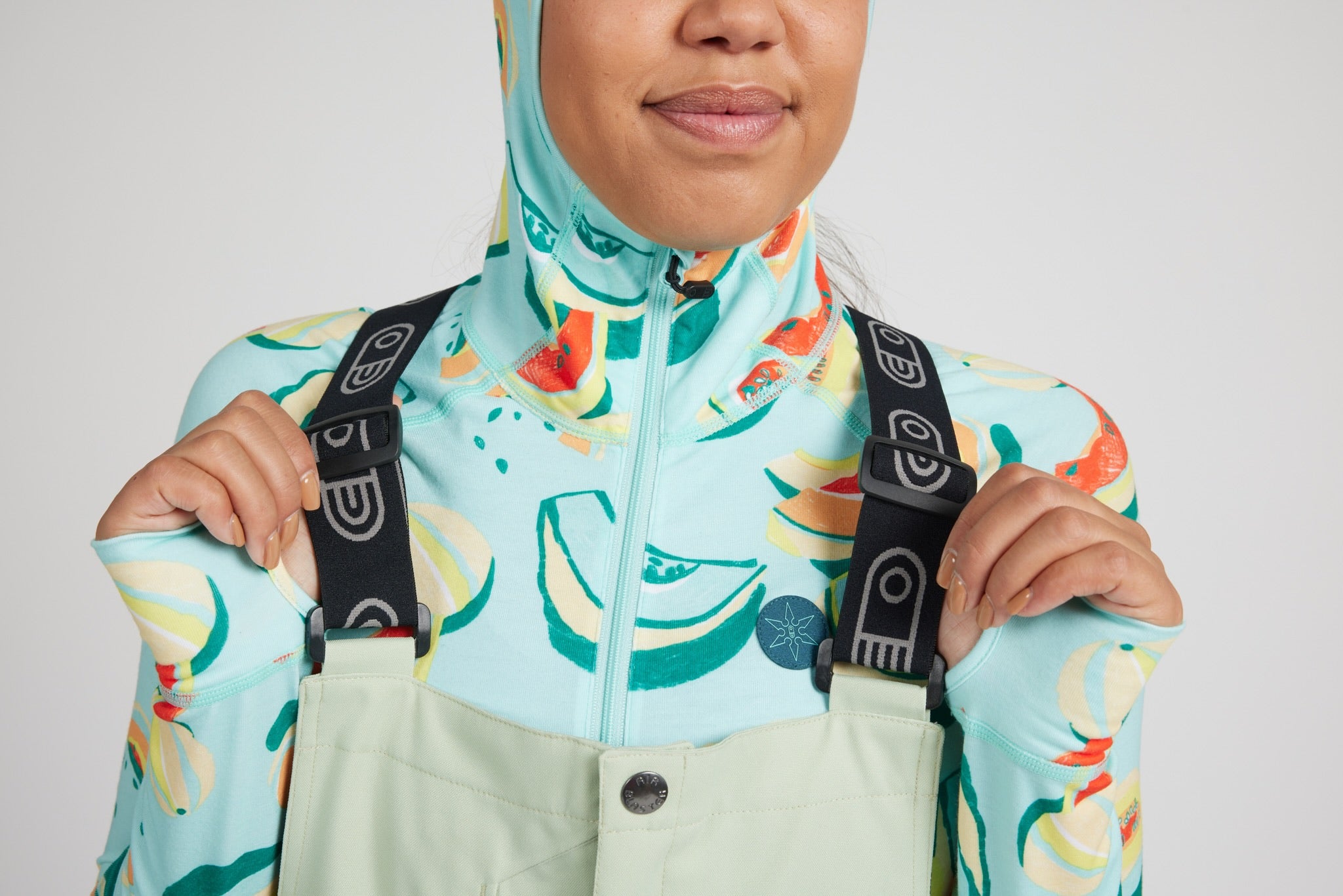 A close-up of a person wearing a light aqua and mint green patterned ski jacket with black adjustable suspenders over sage green bib pants, against a light gray studio background.