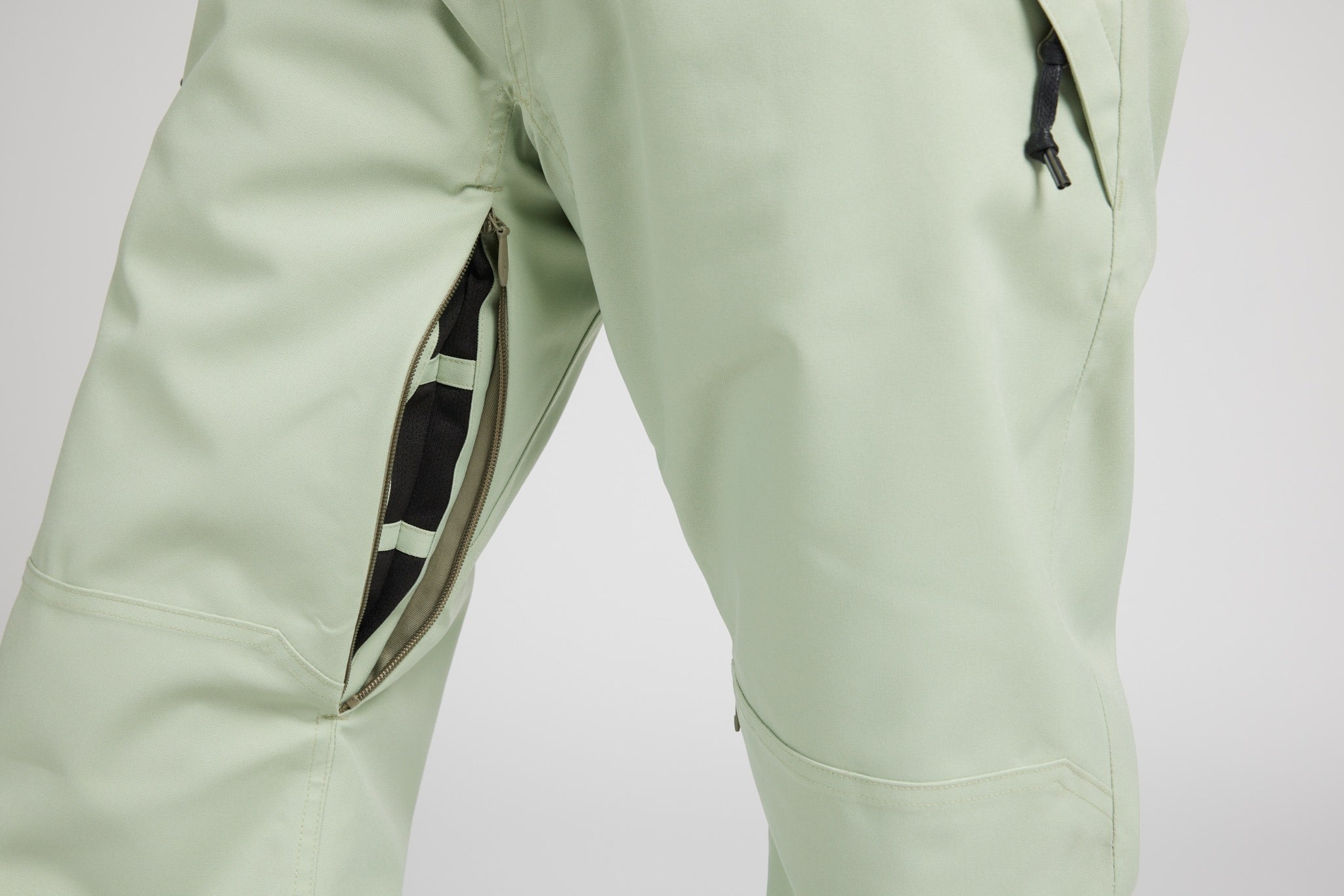 Close-up of light sage green outdoor pants showing a zippered pocket with a black and white striped lining on the left thigh.