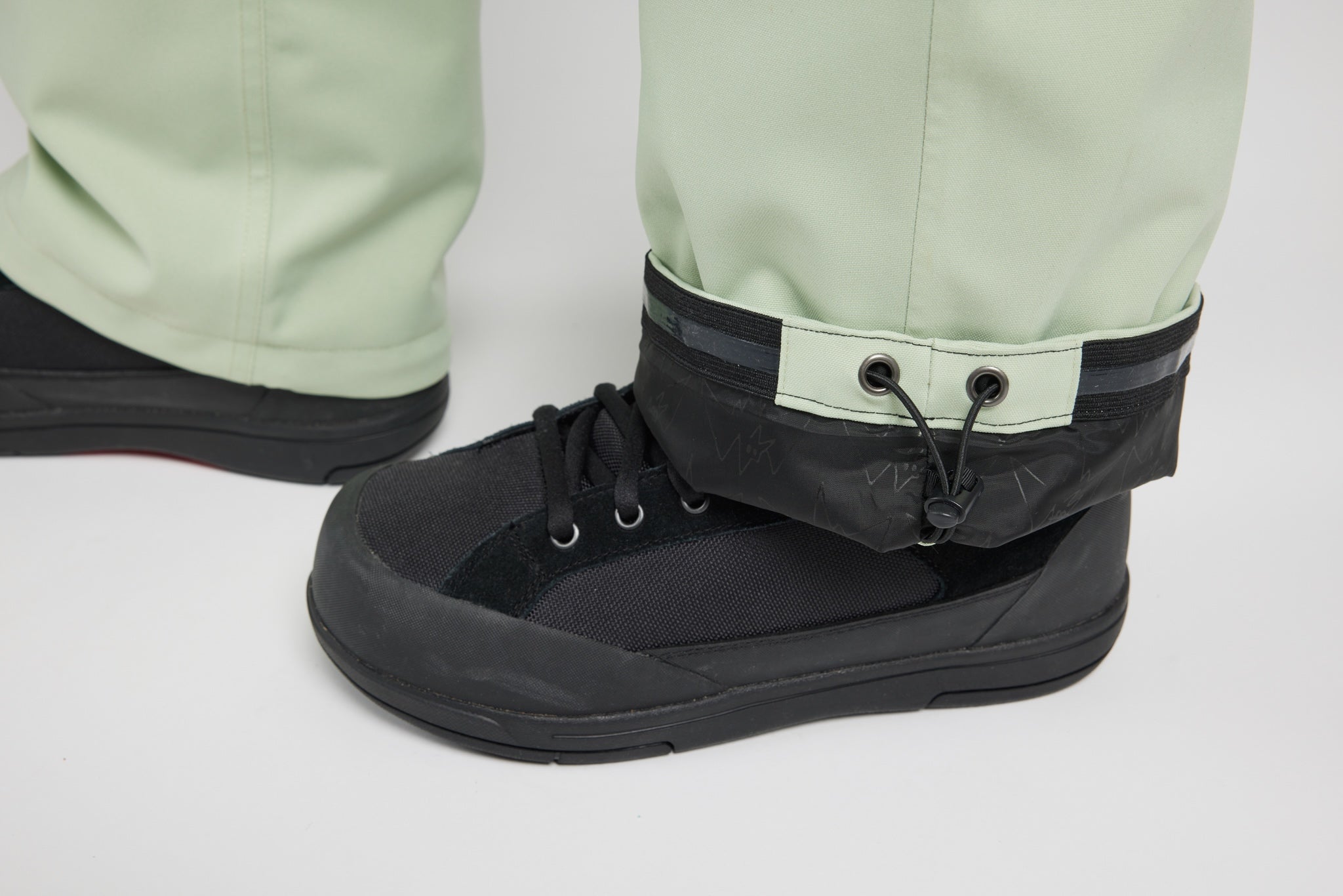Close-up of a person's lower legs wearing light sage green pants and black waterproof ankle boots with a black cuff strap and drawstring.