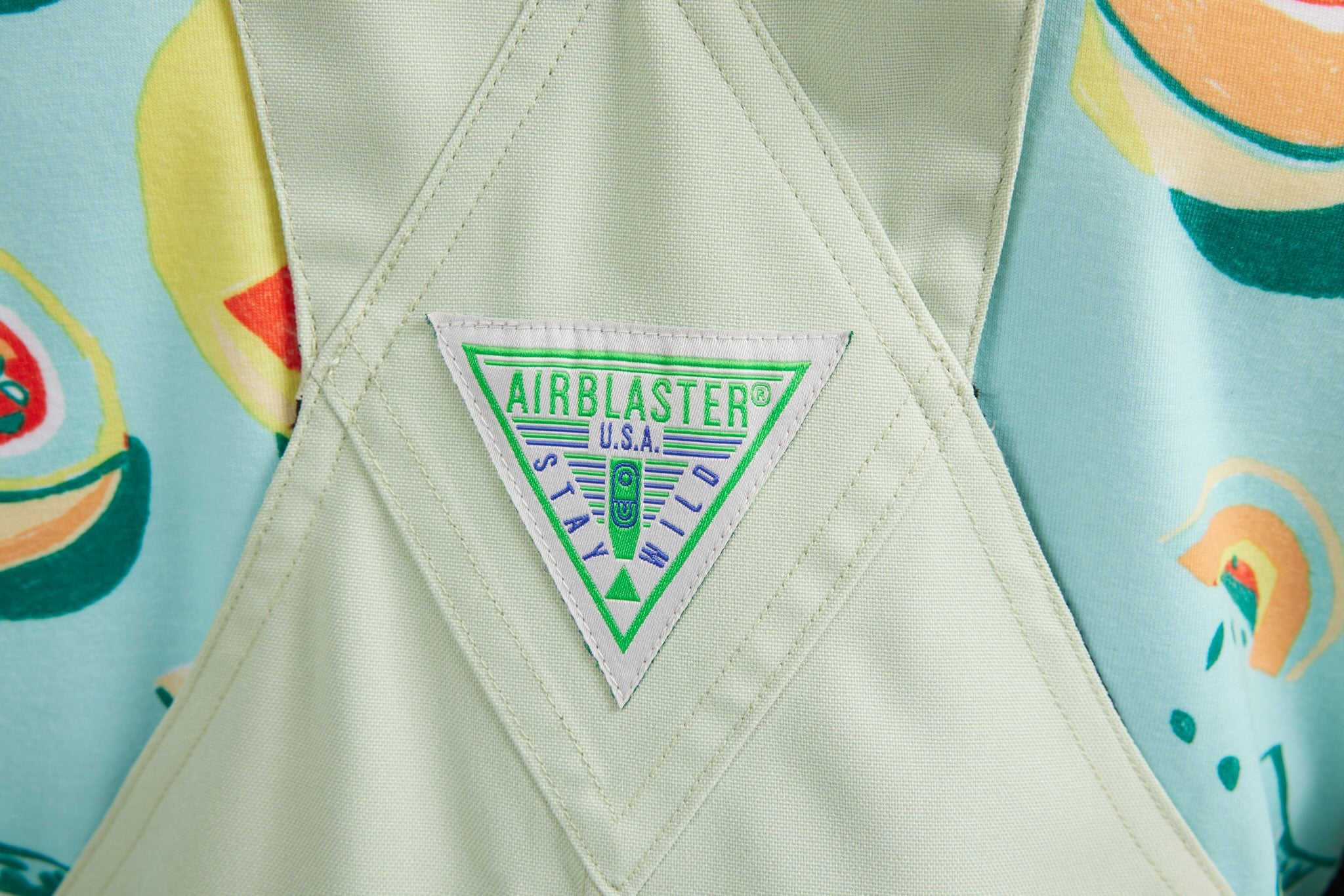 Close-up of a light sage green Airblaster USA 'Stay Wild' patch sewn onto a light green quilted garment, with a colorful avocado-patterned fabric visible in the background.