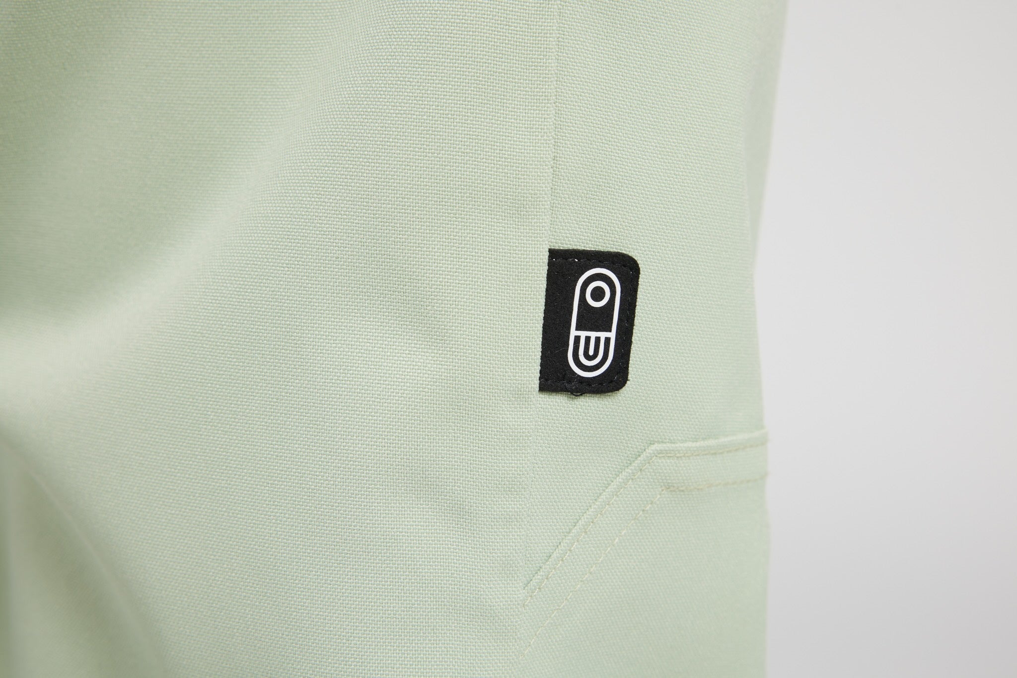 Close-up of a light sage green textured fabric garment with a small black rectangular patch featuring a white stylized 'U' logo, near a pocket seam.