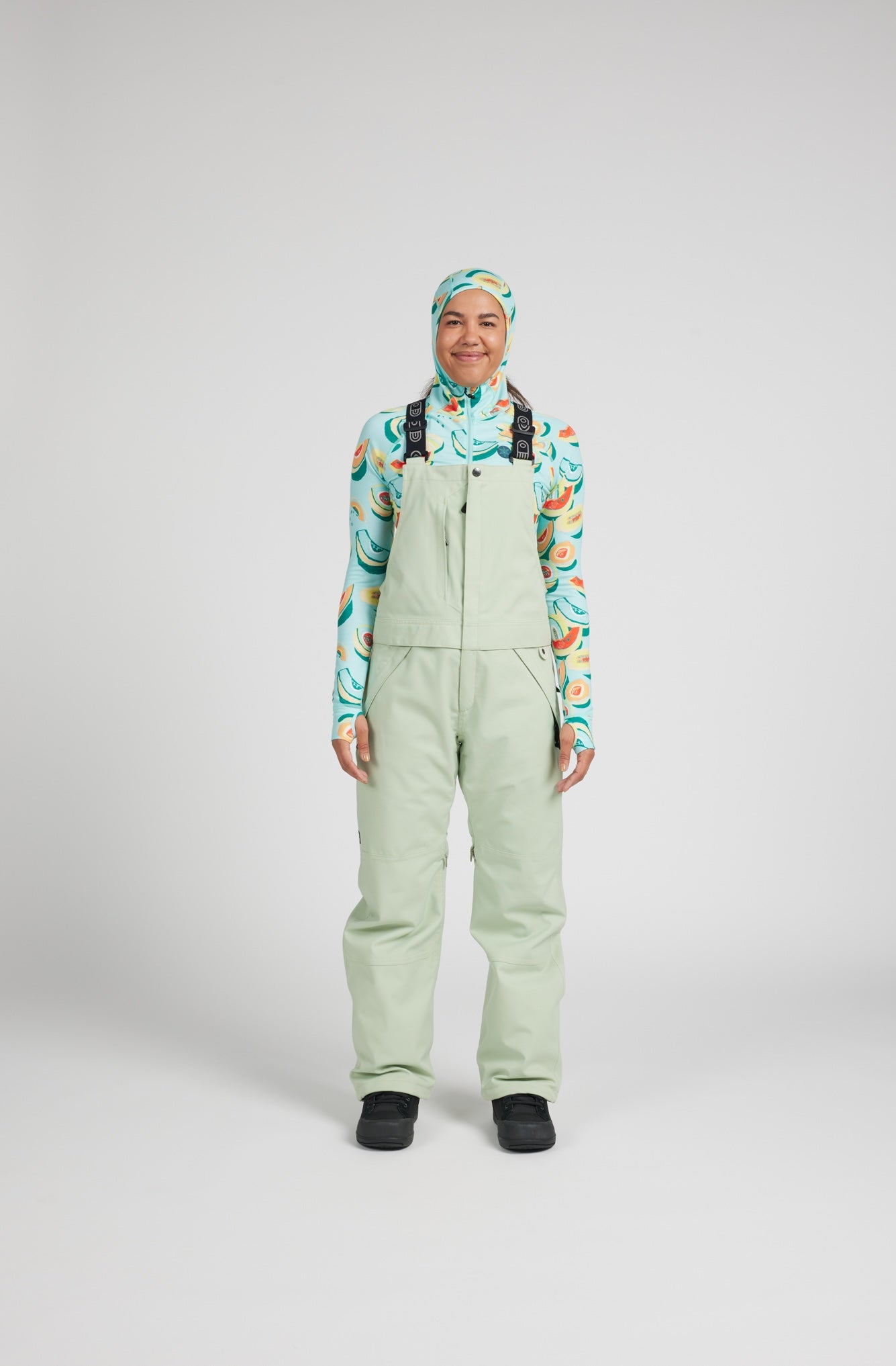 A model stands against a light gray studio background wearing sage green snow bib overalls with black suspenders, a colorful abstract print long-sleeve shirt, and a matching patterned headscarf.