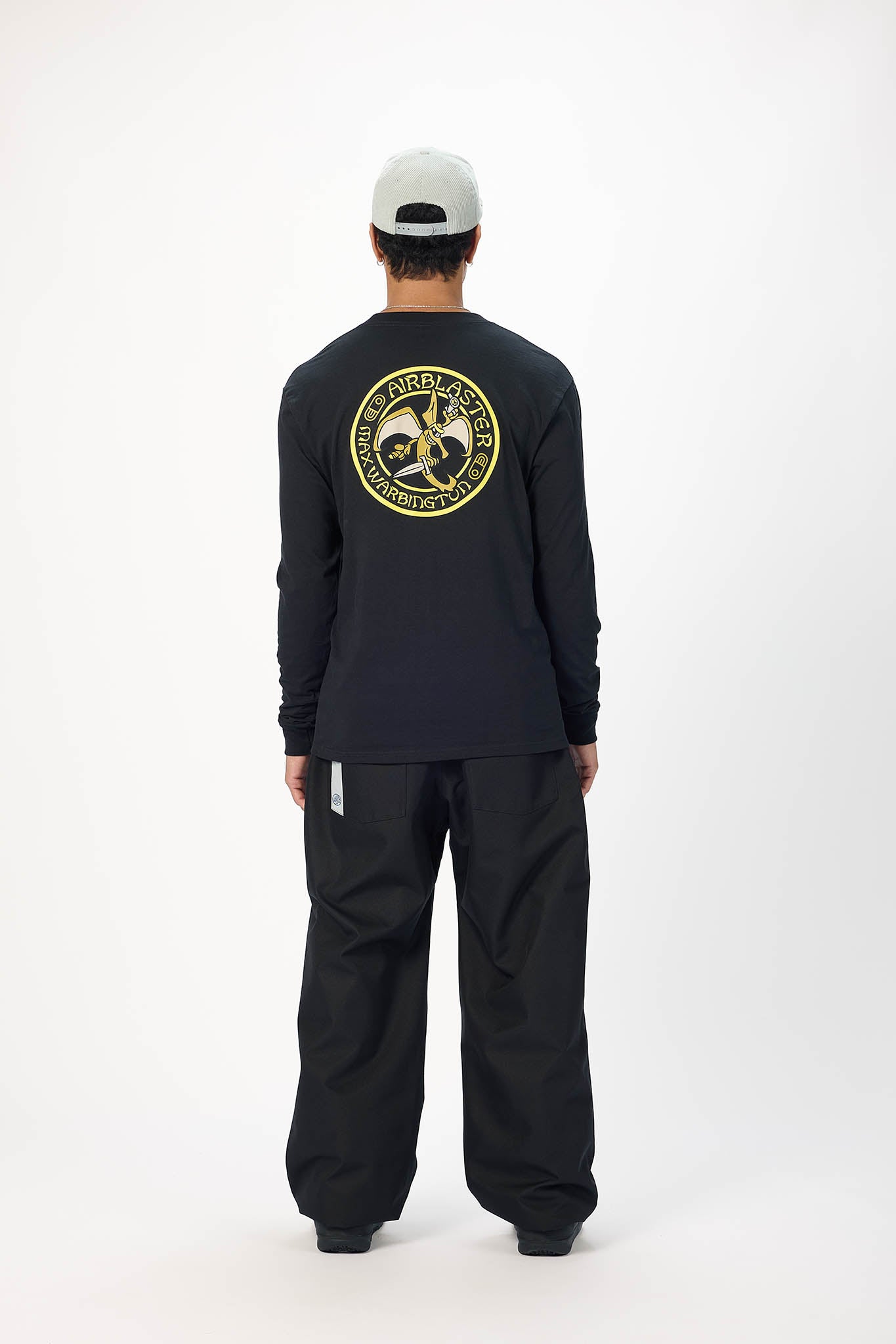 A model shown from the back wearing a black long-sleeve t-shirt with a large circular yellow and white graphic on the back, paired with black pants and a white cap.