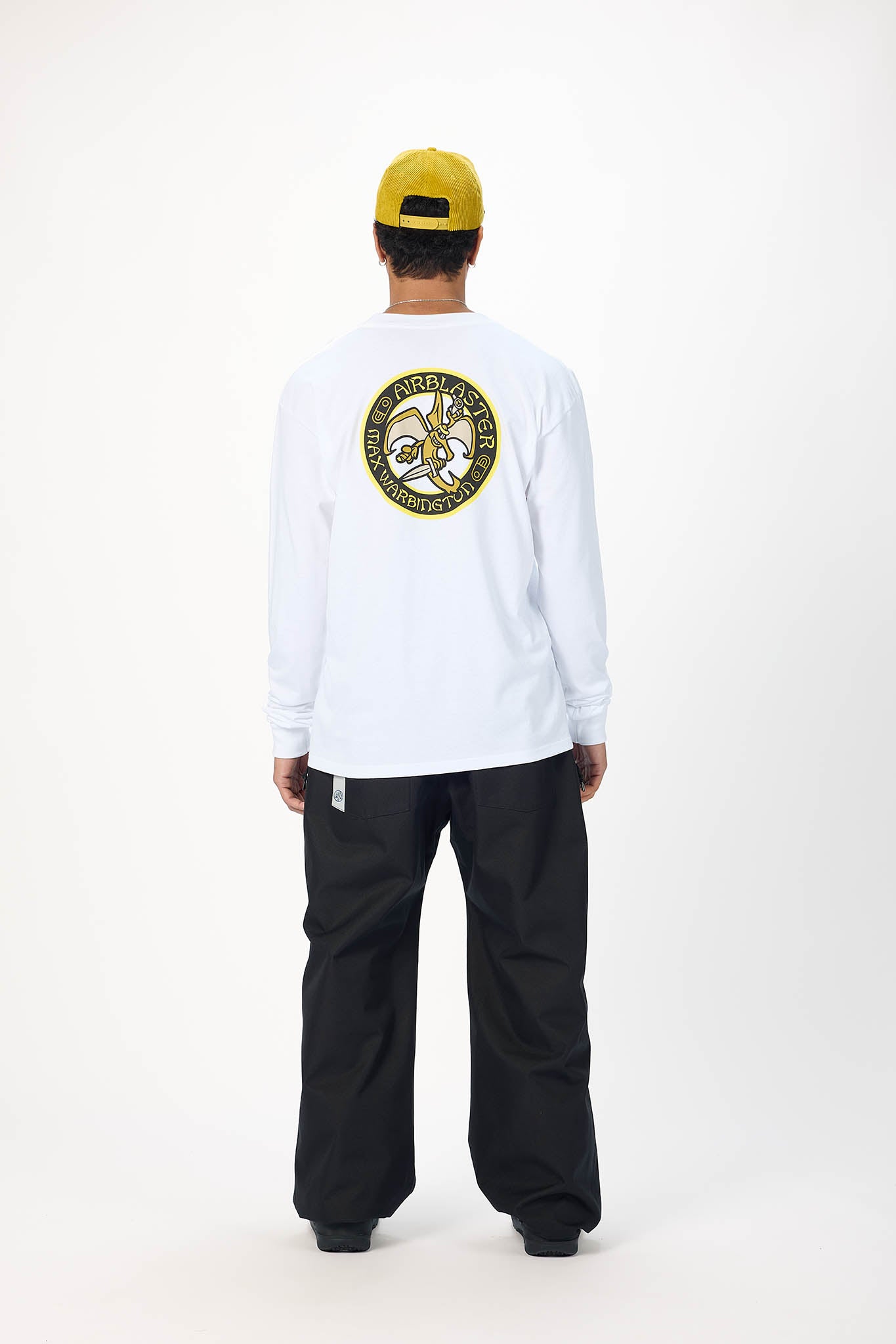 A model shown from the back wearing a white long-sleeve t-shirt with a large circular graphic on the back, black pants, and a yellow baseball cap against a white studio background.