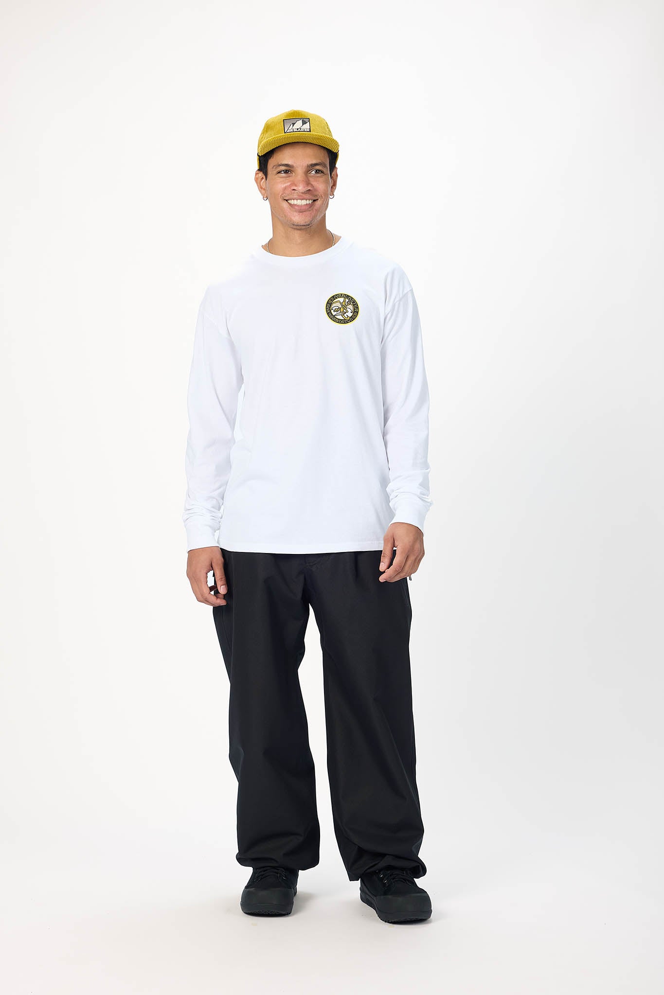 A man modeling a white long-sleeve t-shirt with a circular green and gold logo on the left chest, paired with black pants, black shoes, and a yellow baseball cap, against a plain white studio background.