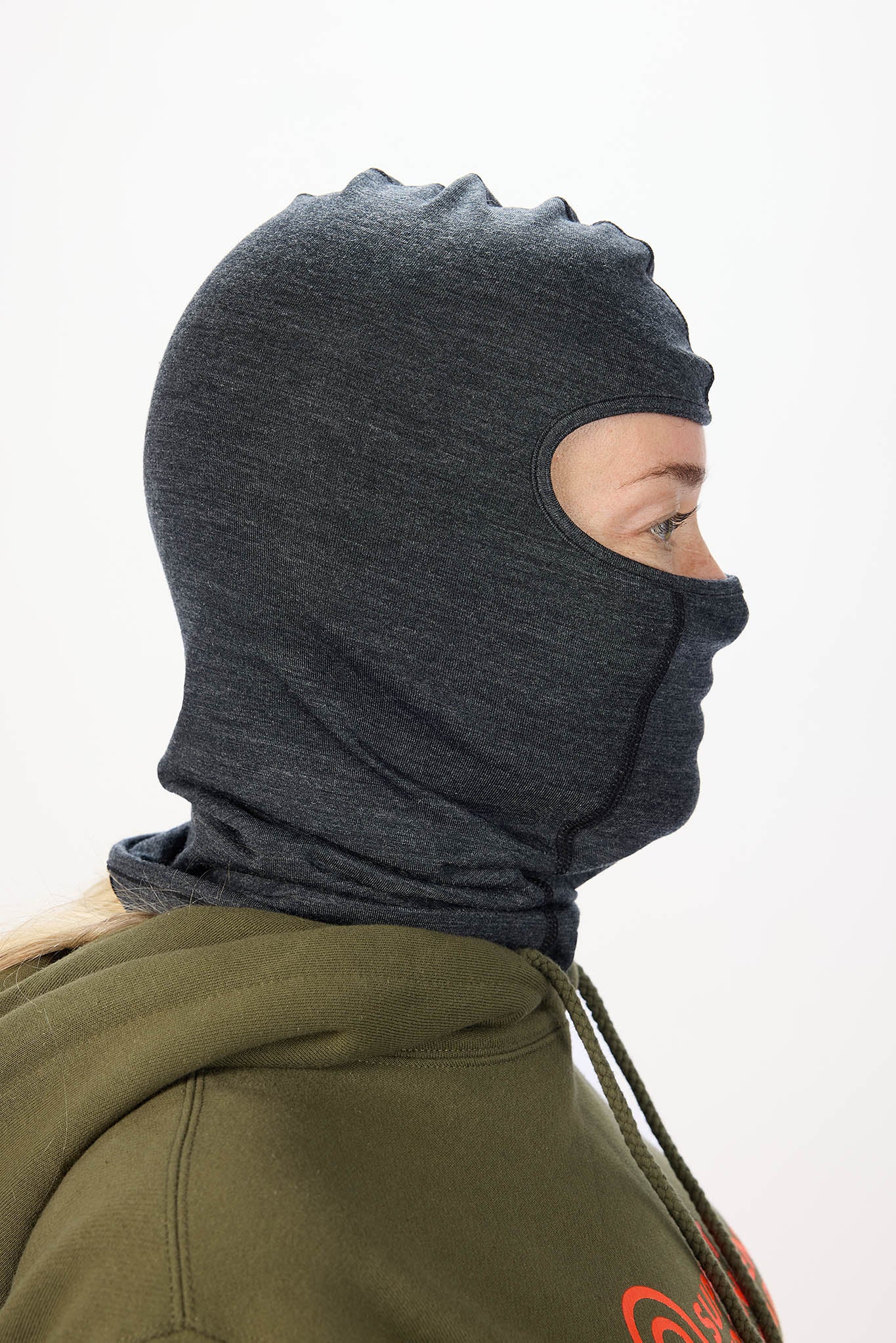 A person shown in side profile wearing a dark heather gray full-head balaclava mask over an olive green hoodie against a plain white background.