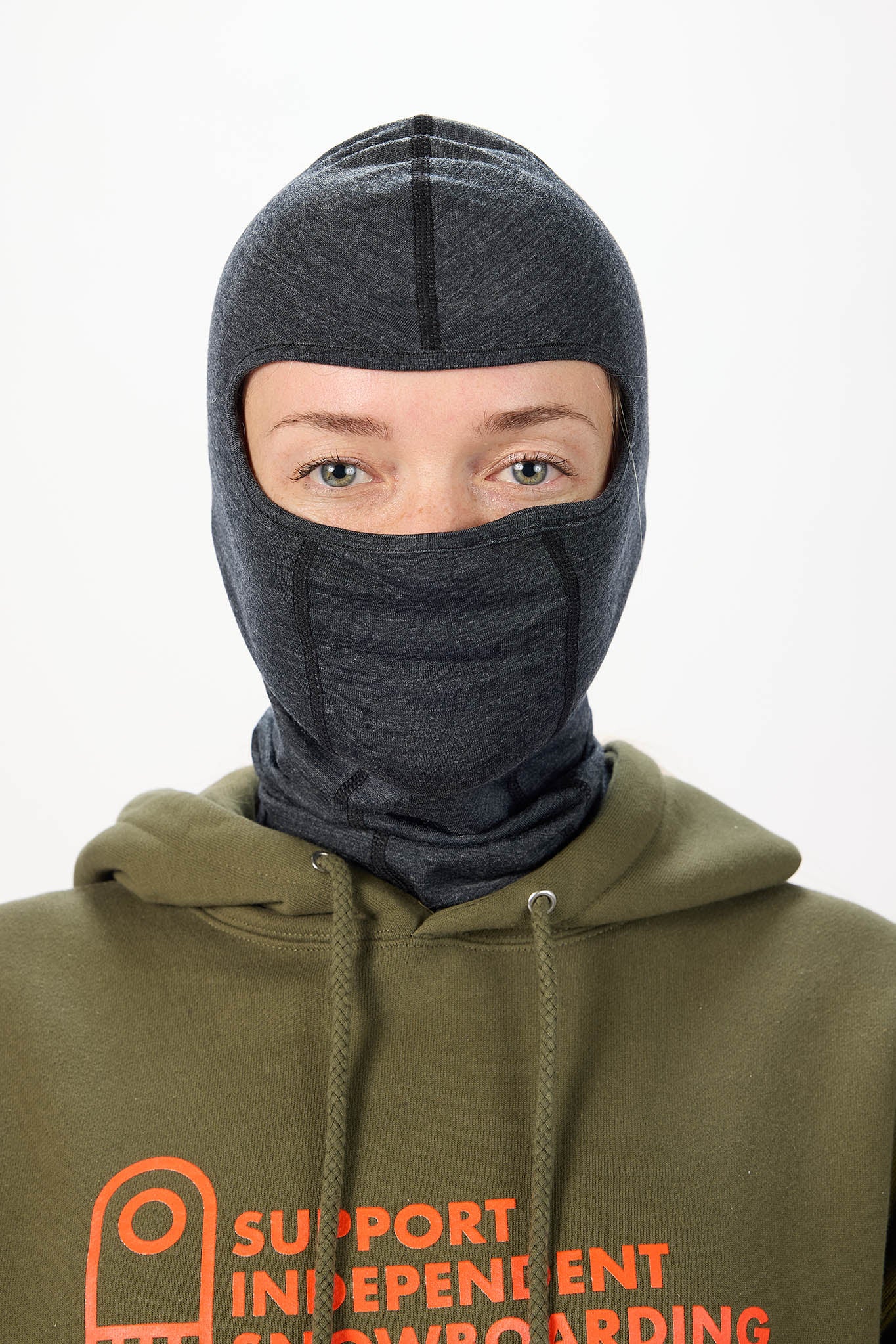 A person wearing a dark gray heathered ski balaclava mask over their face, paired with an olive green hoodie featuring orange text that reads 'SUPPORT INDEPENDENT SNOWBOARDING'.
