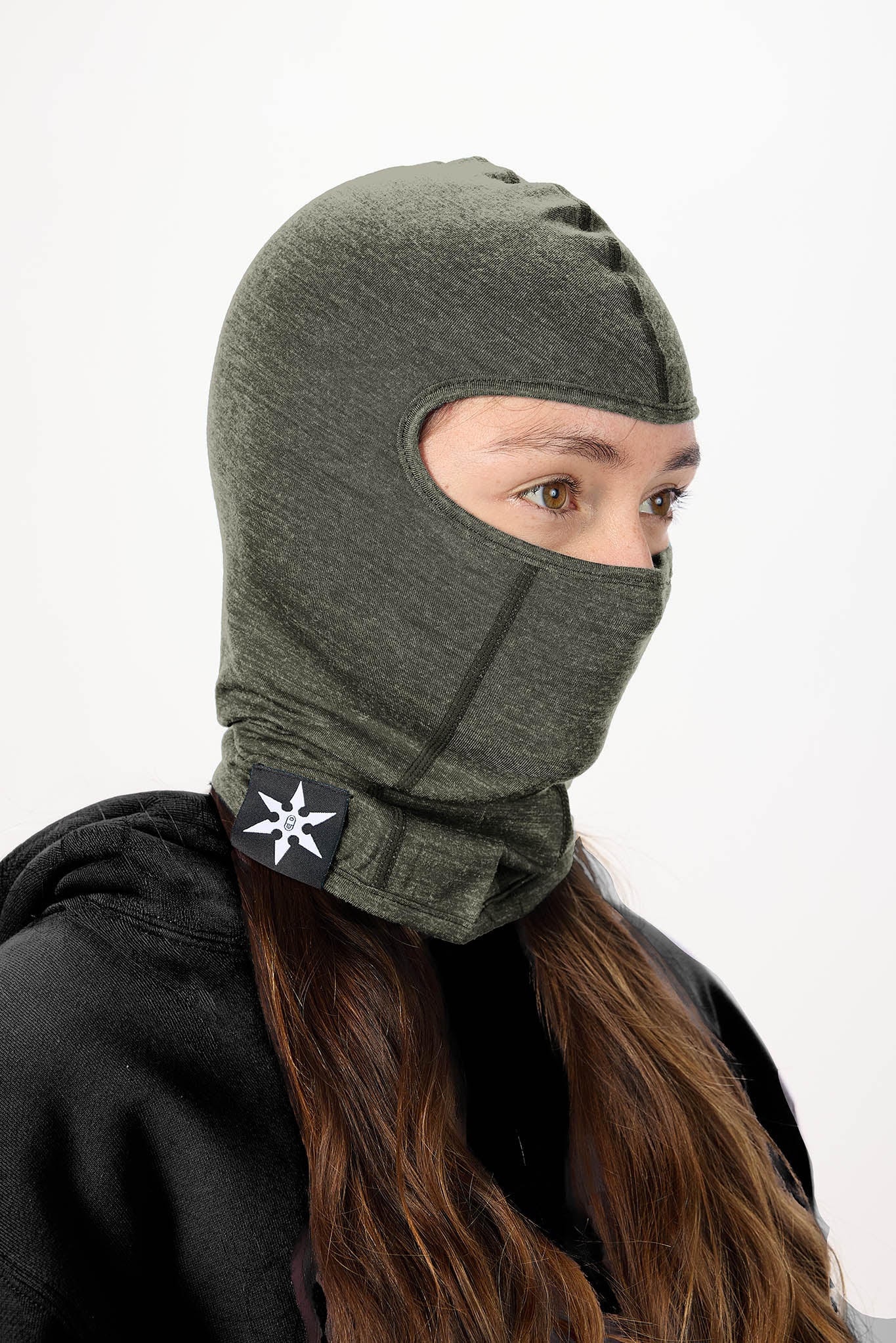 A woman wearing an olive green knit balaclava mask with a black chin strap featuring a white geometric logo, set against a plain white background.