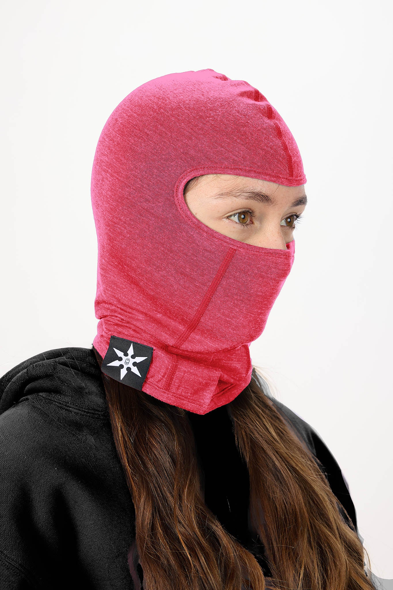 A person wearing a bright pink balaclava face covering with a black strap and white logo patch, paired with a black hoodie against a plain white background.