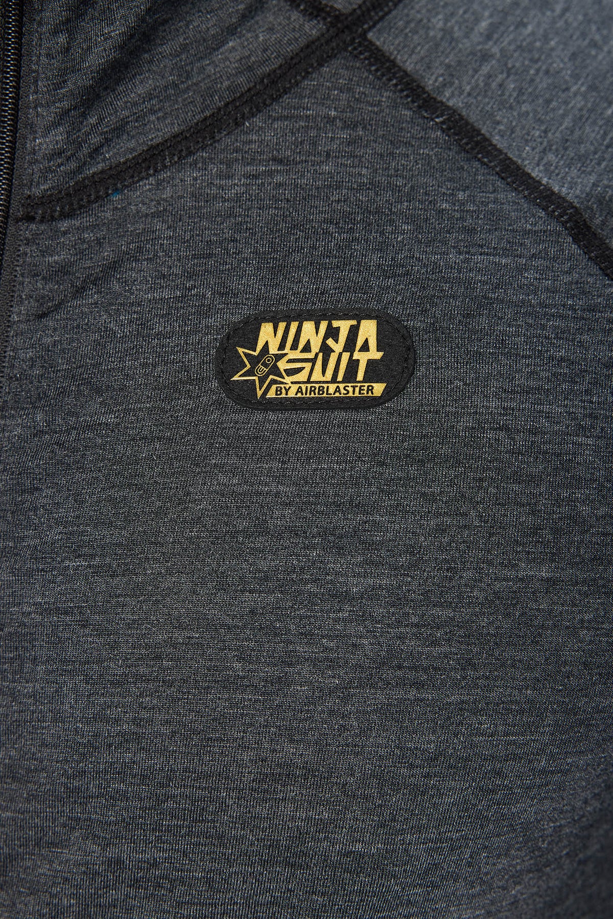 Close-up of a dark heather gray athletic jacket with a black oval patch on the chest that reads 'NINJA SUIT' and 'BY AERBLASTER' in gold lettering.