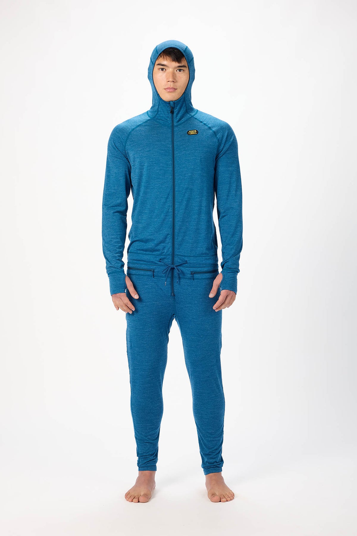 A man wearing a ocean blue hooded zip-up onesie , standing barefoot against a plain white studio background.