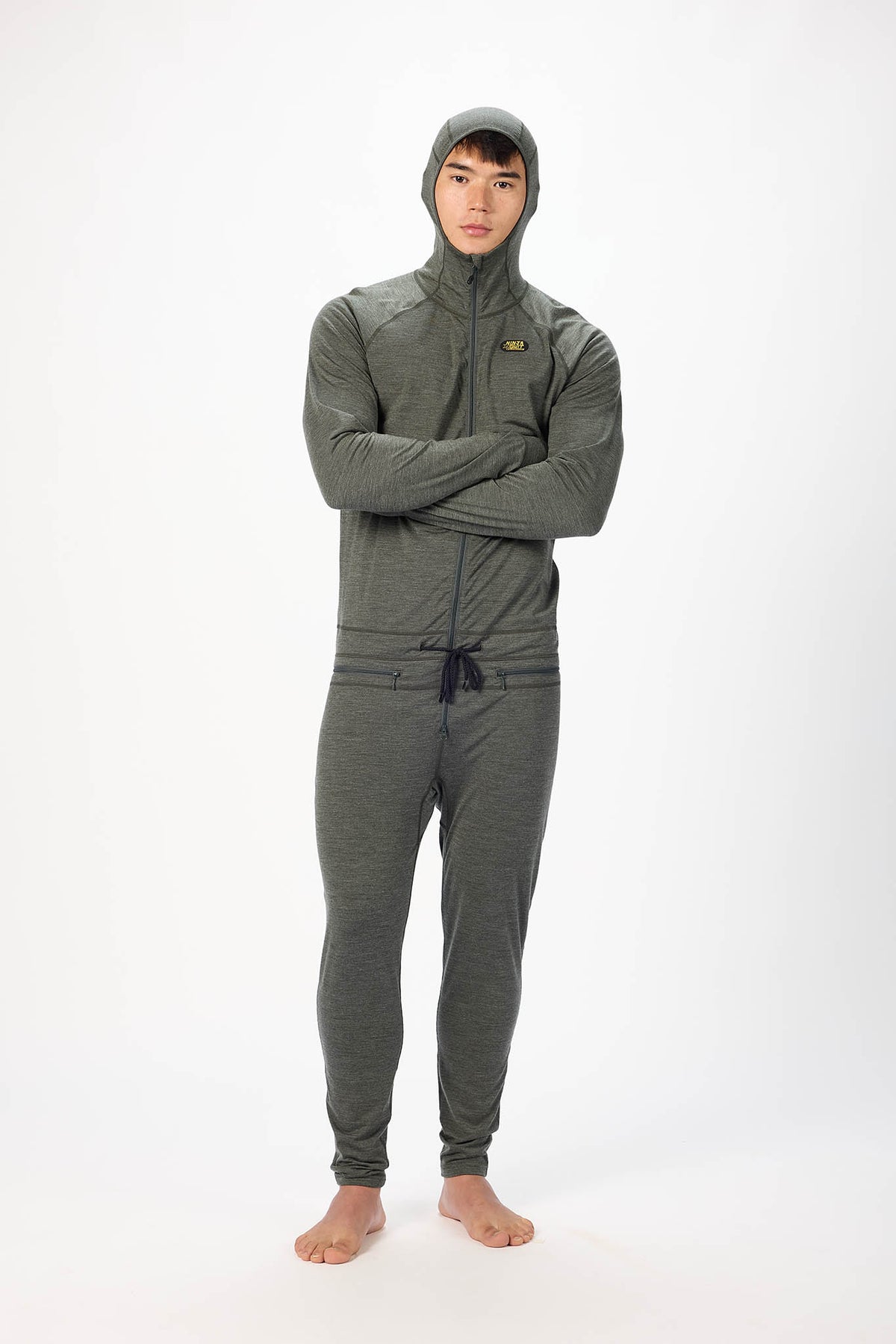A man wearing a dark gray hooded zip-up onesie , standing barefoot against a plain white studio background.