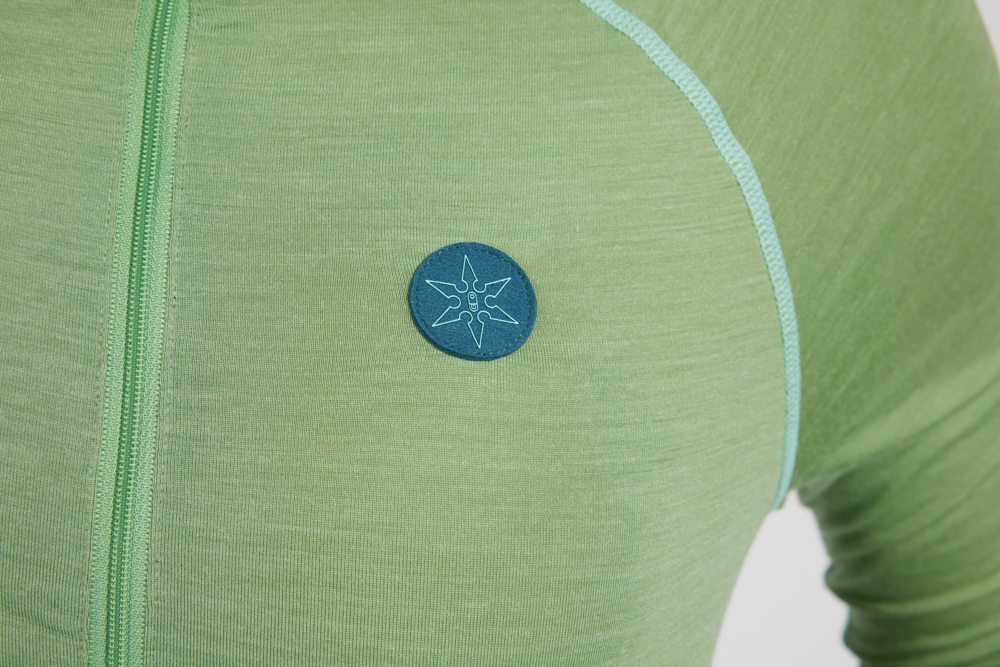 Close-up of a light green long-sleeve athletic top with a teal circular logo patch featuring a compass rose design on the chest.