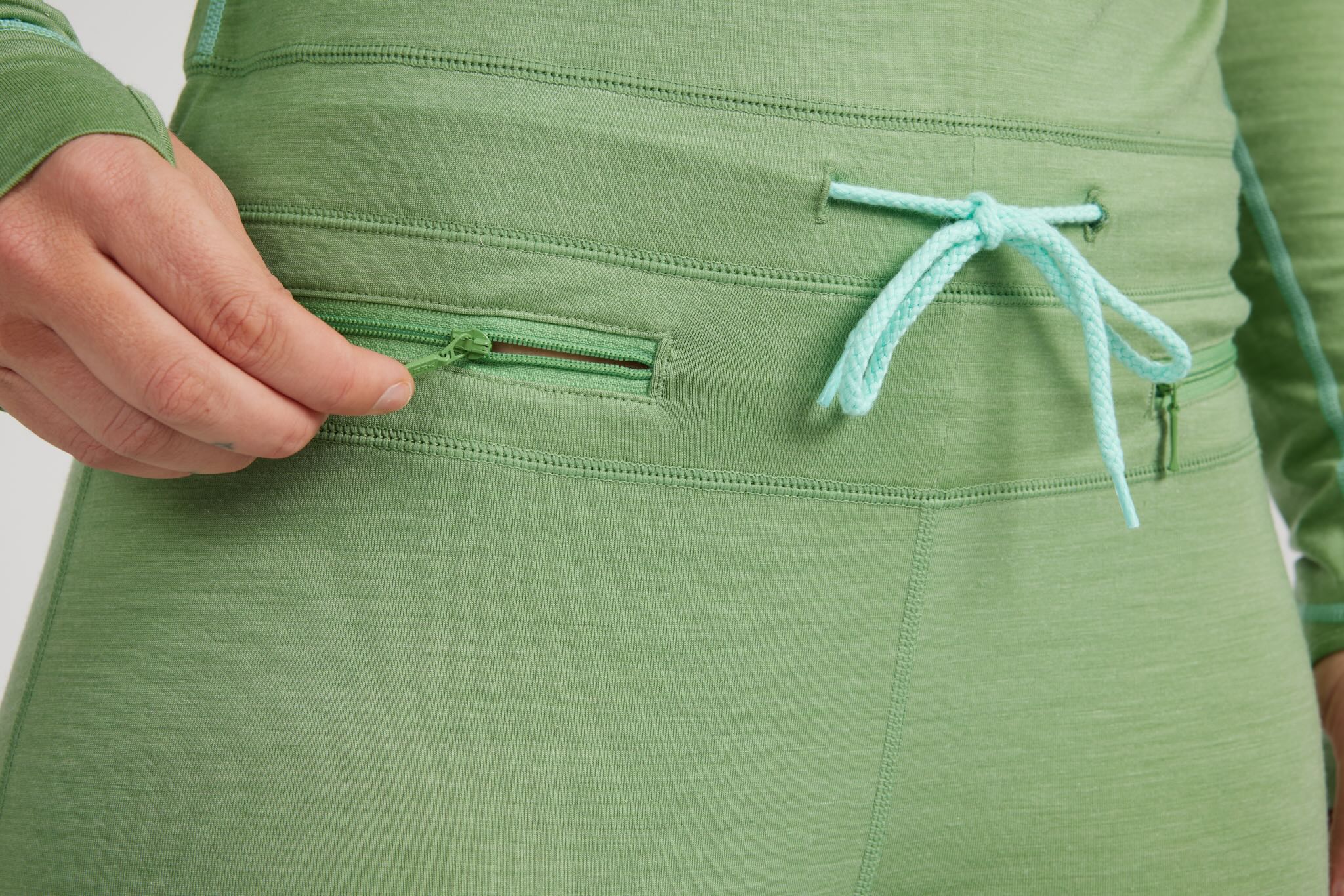 Close-up of a person wearing a sage green athletic top, showing a zippered pocket being opened with a hand and a mint green drawstring at the waist.