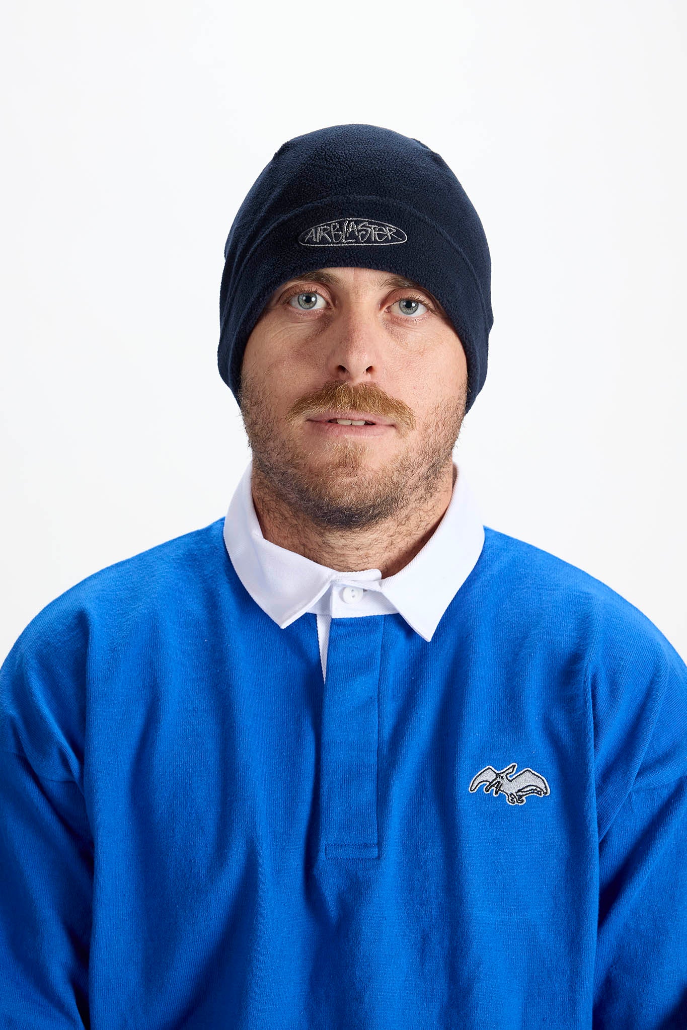 A man wearing a black beanie with a white embroidered logo and a blue rugby-style polo shirt with a white collar, photographed against a plain white background.