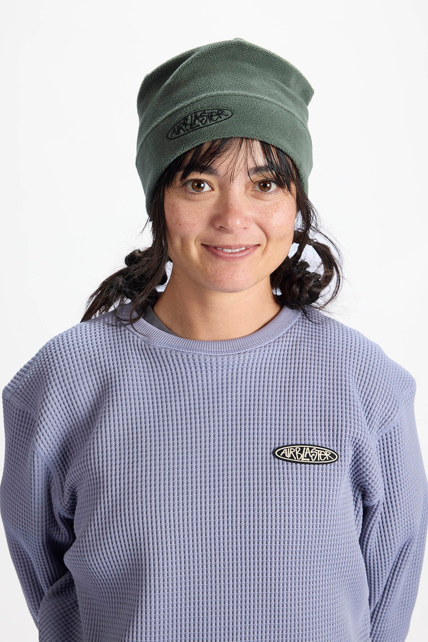 A woman wearing a muted green beanie and a lavender-purple waffle-knit sweatshirt, both featuring an oval black and white 'ANY KNOT' logo on the front.