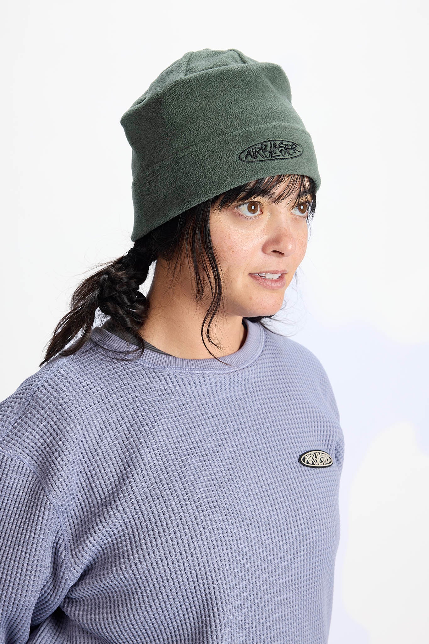 A woman wearing an olive green beanie with a black embroidered logo and a matching waffle-knit sweatshirt, photographed against a plain white background.