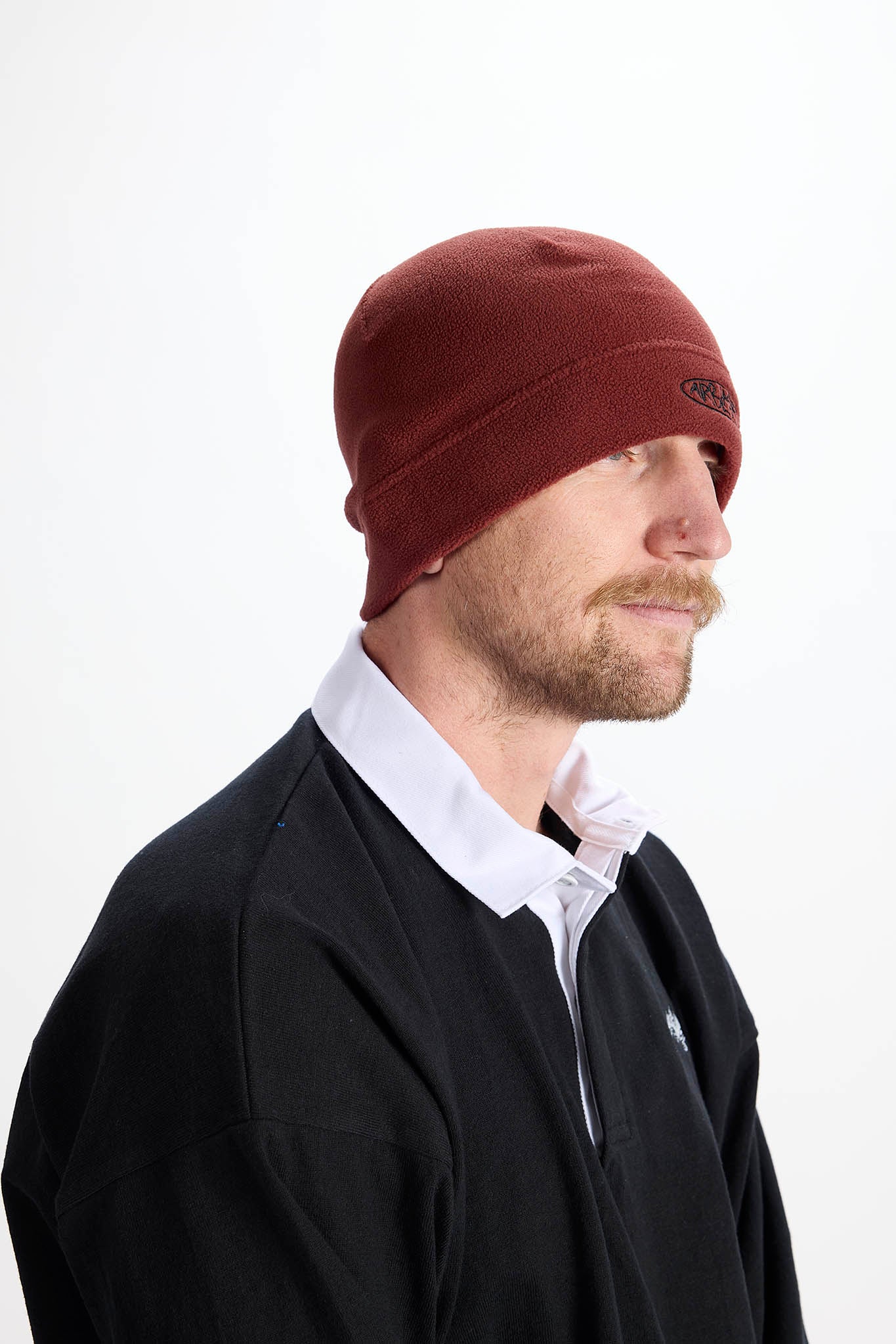 A man in profile wearing a dark red knit beanie, a black zip-up sweater, and a white collared shirt, photographed against a plain white studio background.