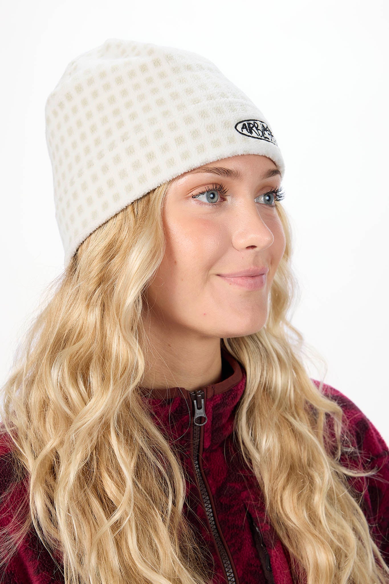A woman with long blonde hair wearing a cream-colored beanie with a small black logo on the side, paired with a burgundy fleece jacket, against a plain white background.