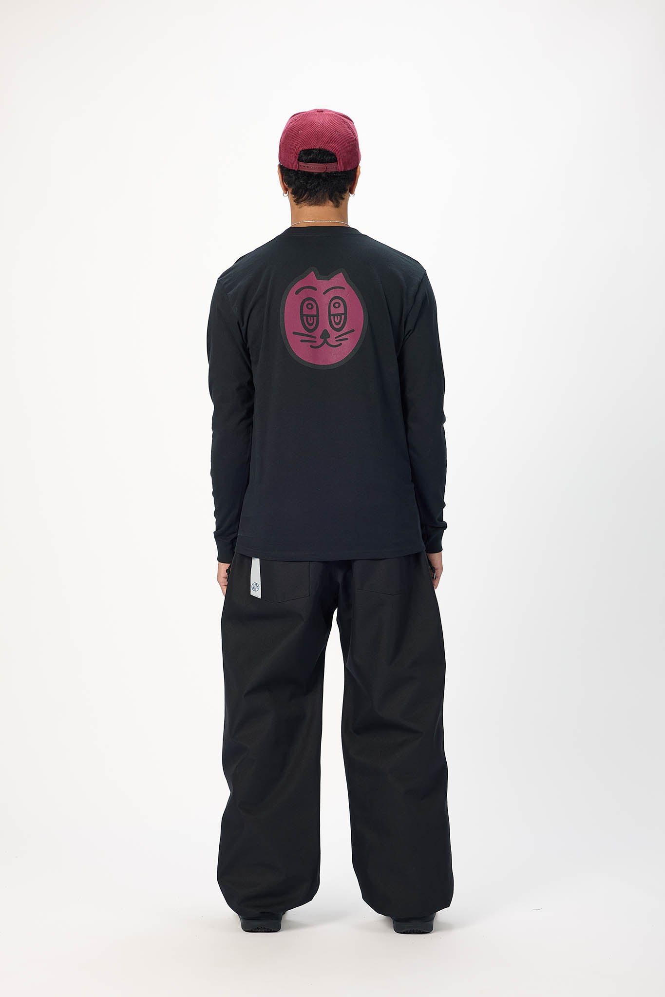 A person shown from the back wearing a black long-sleeve shirt with a pink graphic on the back, black pants, and a burgundy cap, photographed against a plain light gray studio background.