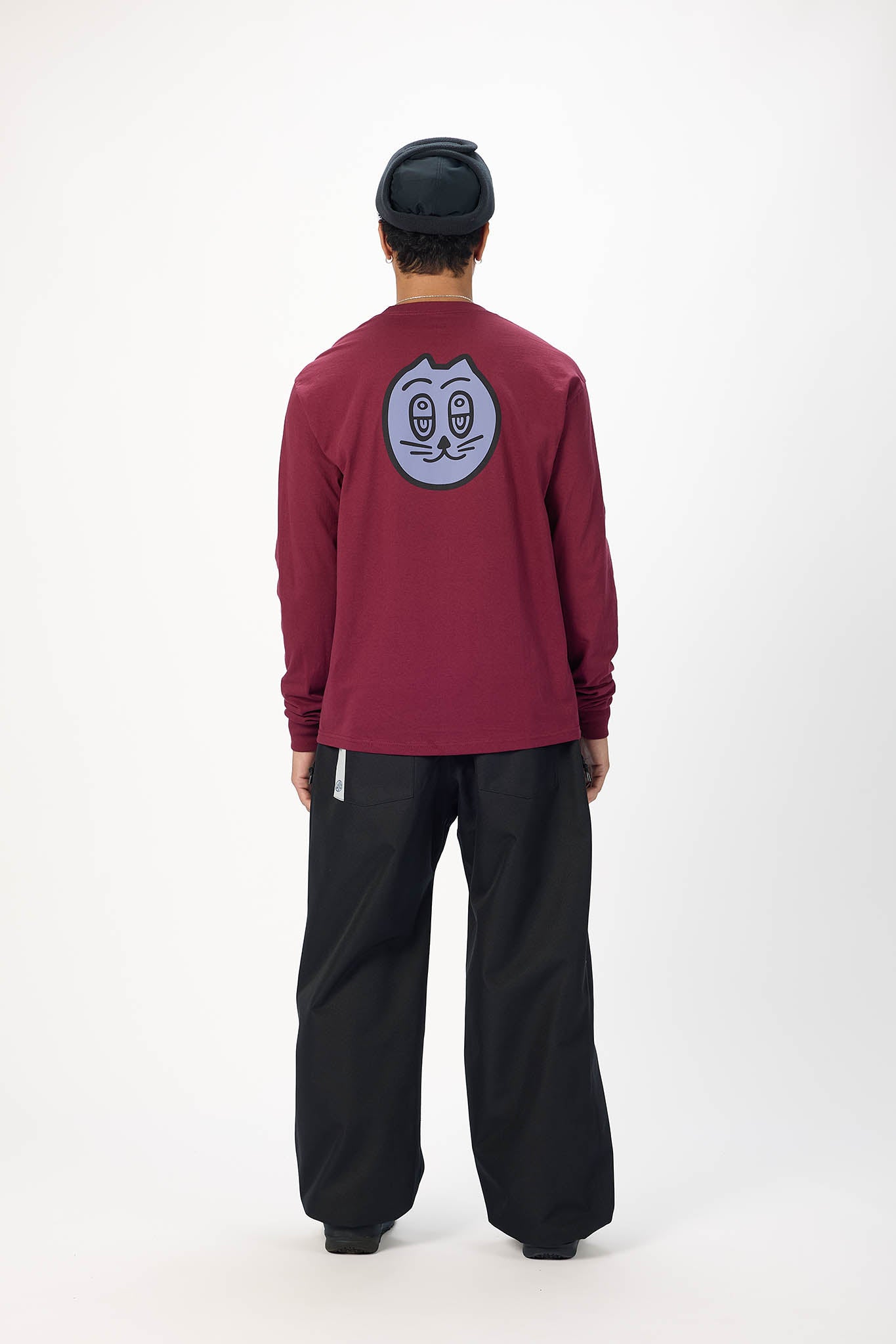 A person shown from the back wearing a burgundy long-sleeve shirt with a circular graphic on the upper back, black pants, and a black beanie, photographed against a plain light gray studio background.