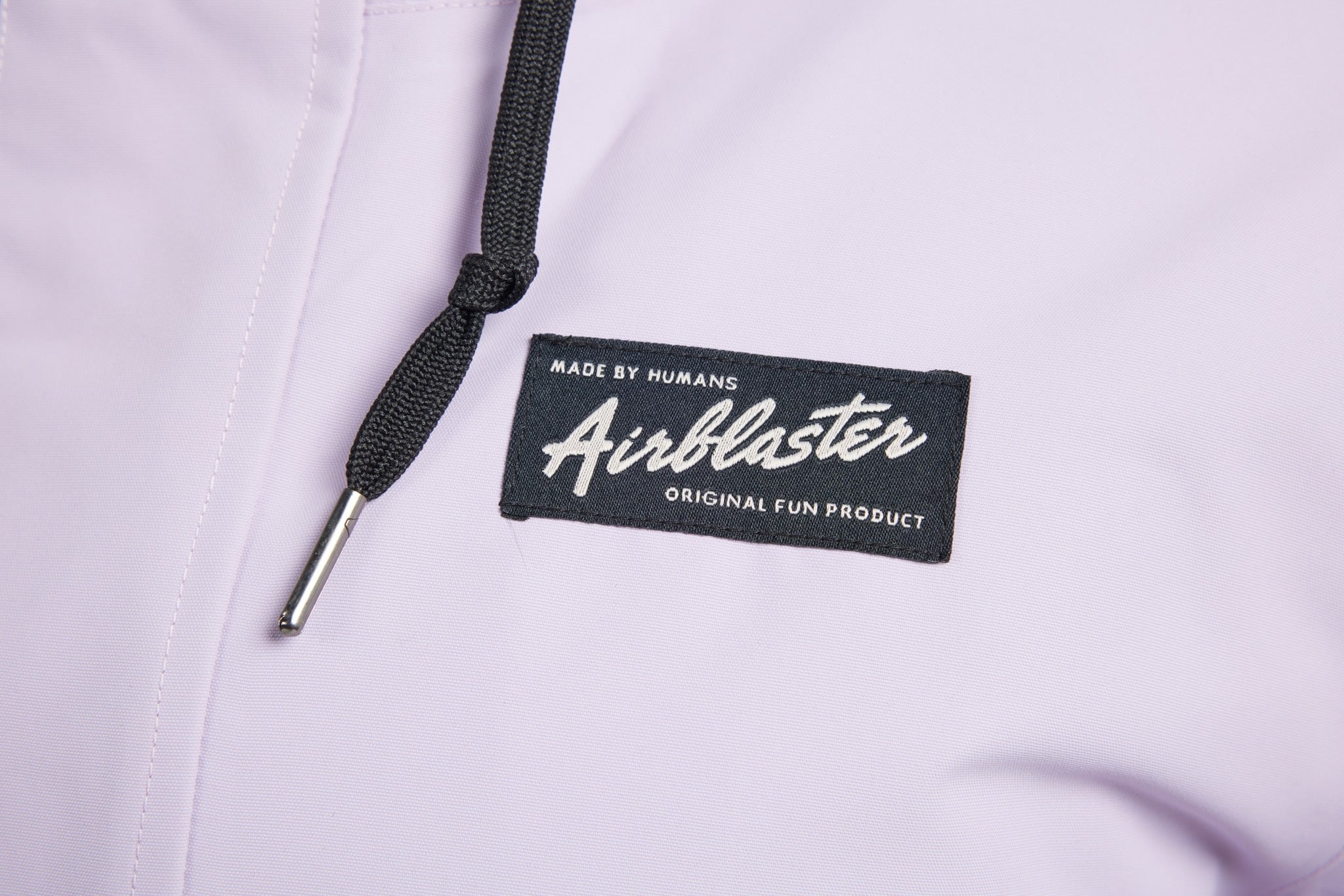 Close-up of a light lavender or pale purple garment with a black drawstring and a black fabric brand patch reading 'Airblaster' and 'Made by Humans Original Fun Product'.