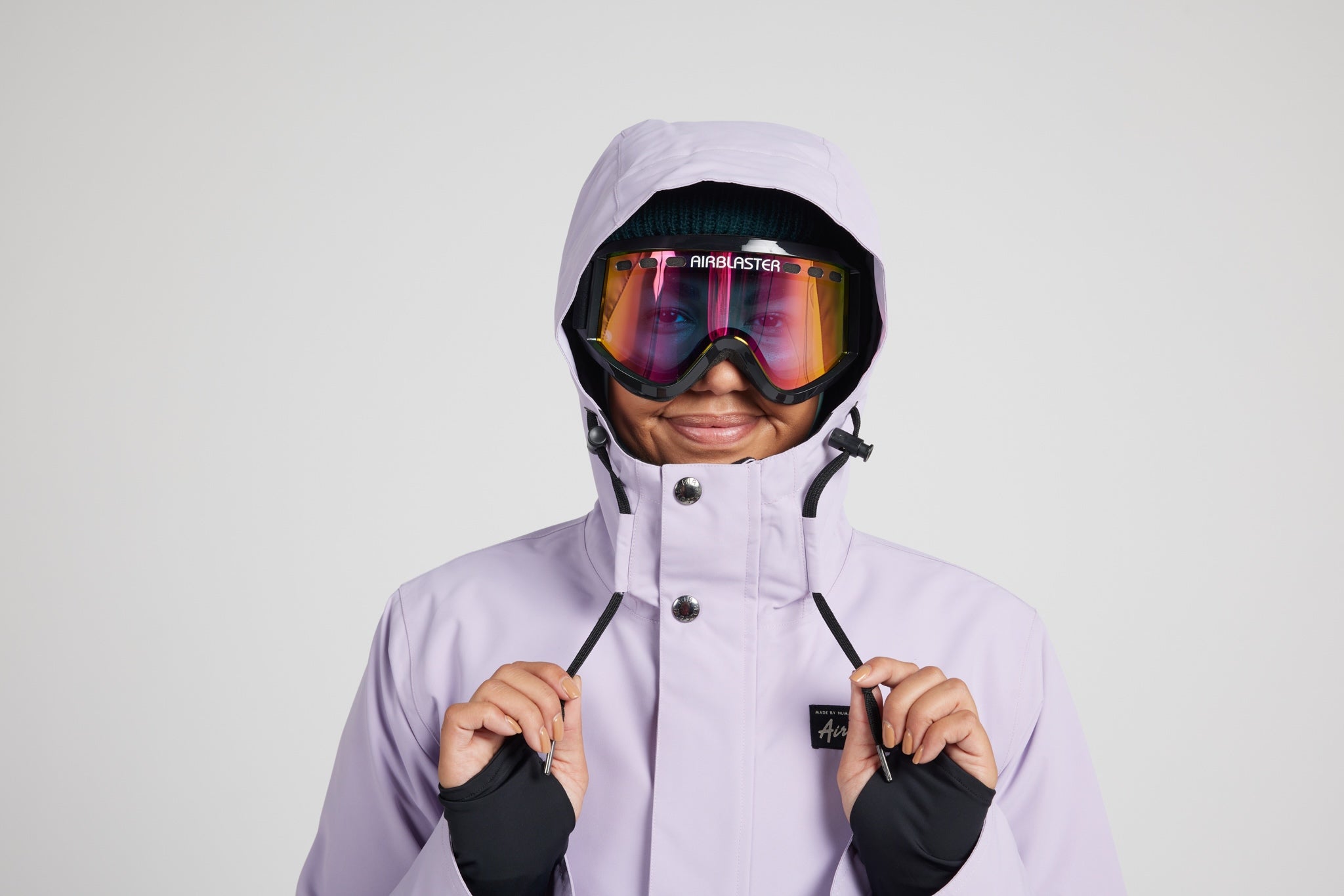 A model wearing a light lavender ski jacket, black gloves, and ski goggles with a colorful reflective lens. The jacket features a high collar with snap buttons and a small black logo patch on the chest, set against a solid light gray background.