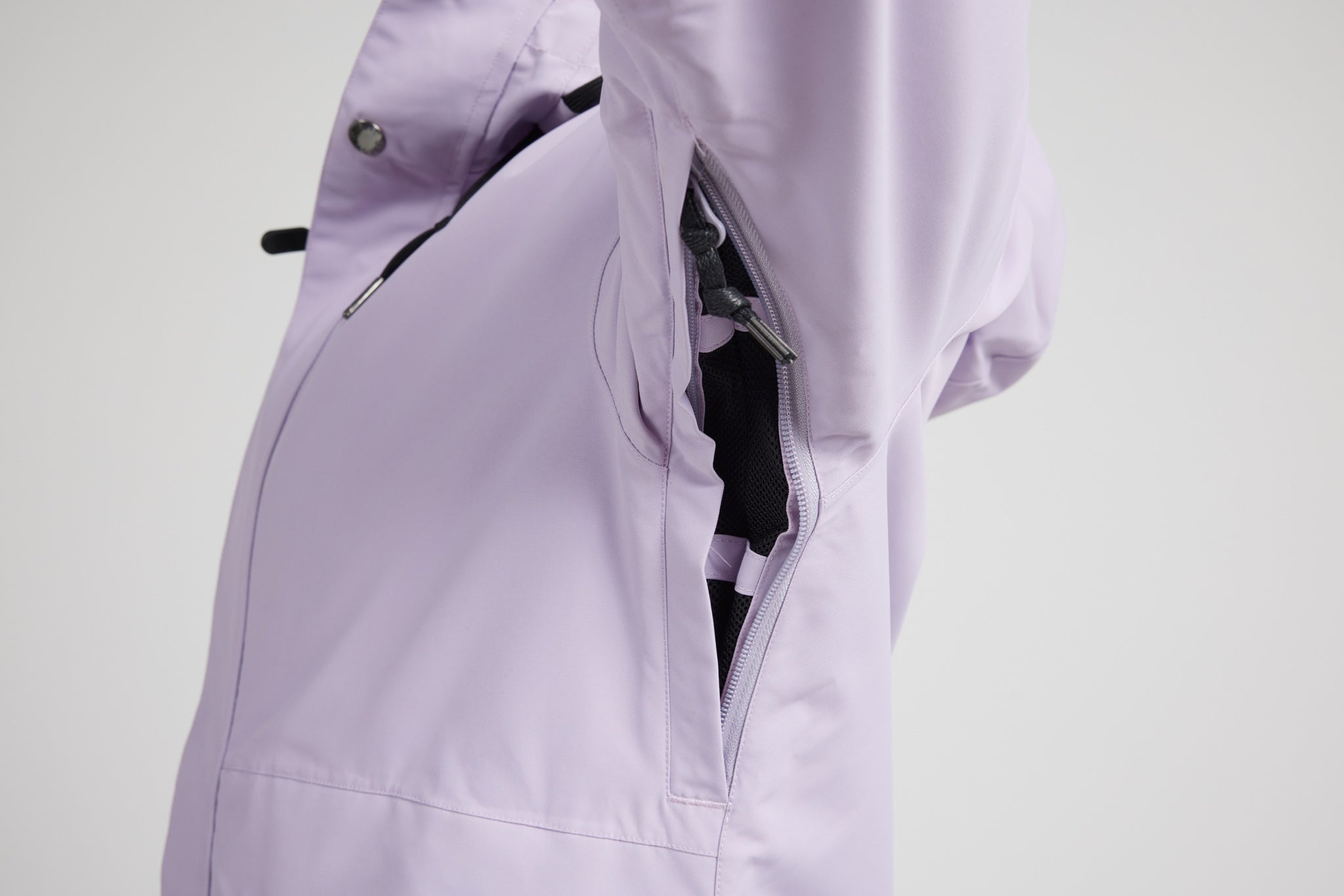 Close-up of a light lavender technical jacket with a side zip pocket open, showing black interior lining and zipper details against a light gray background.