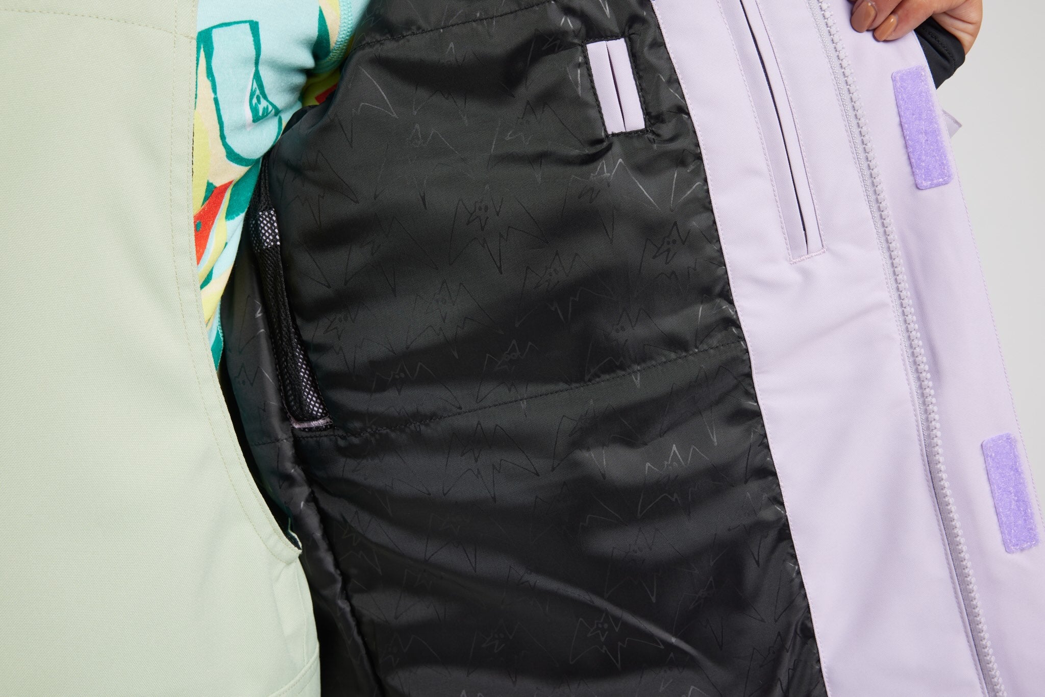 Close-up detail of a ski jacket showing the black patterned inner shell, a light lavender outer panel with a zipper, and a small portion of a multicolor graphic fabric.