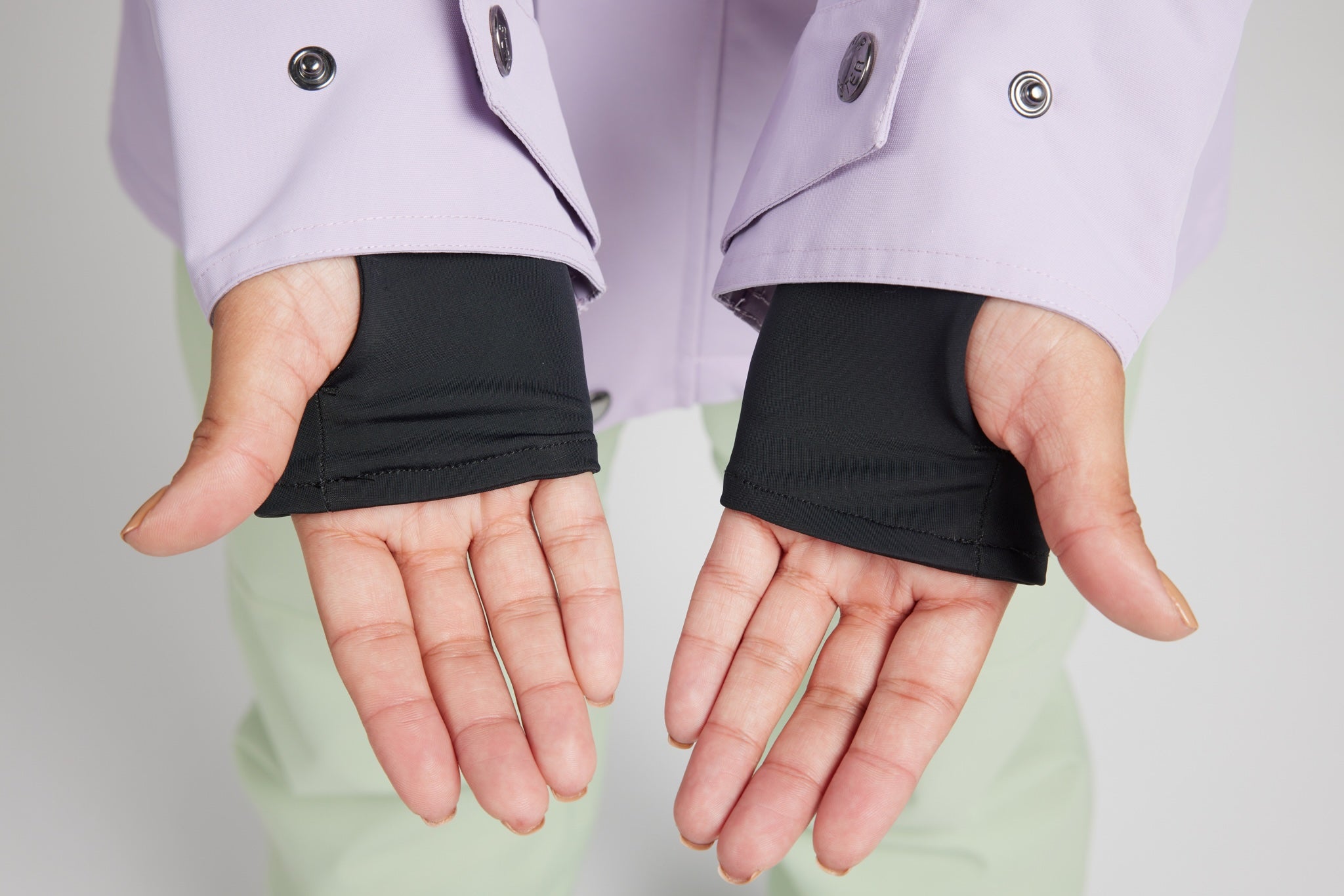 Close-up of a person's hands wearing black fingerless sleeve gloves under a light purple jacket, with pale green pants visible in the background.