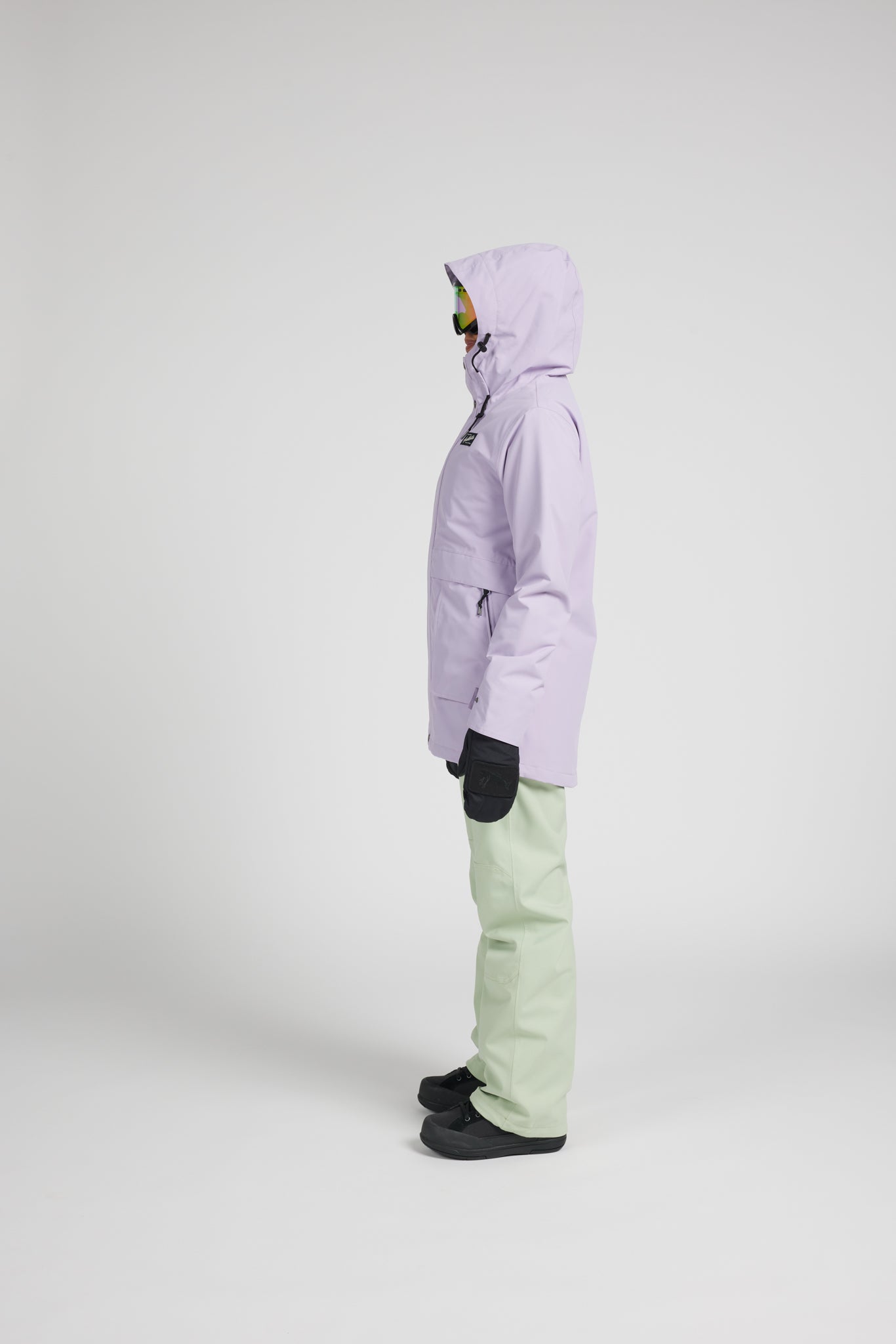 A side-profile studio shot of a person wearing a light lavender hooded ski jacket, sage green snow pants, black gloves, and black shoes against a light gray background.